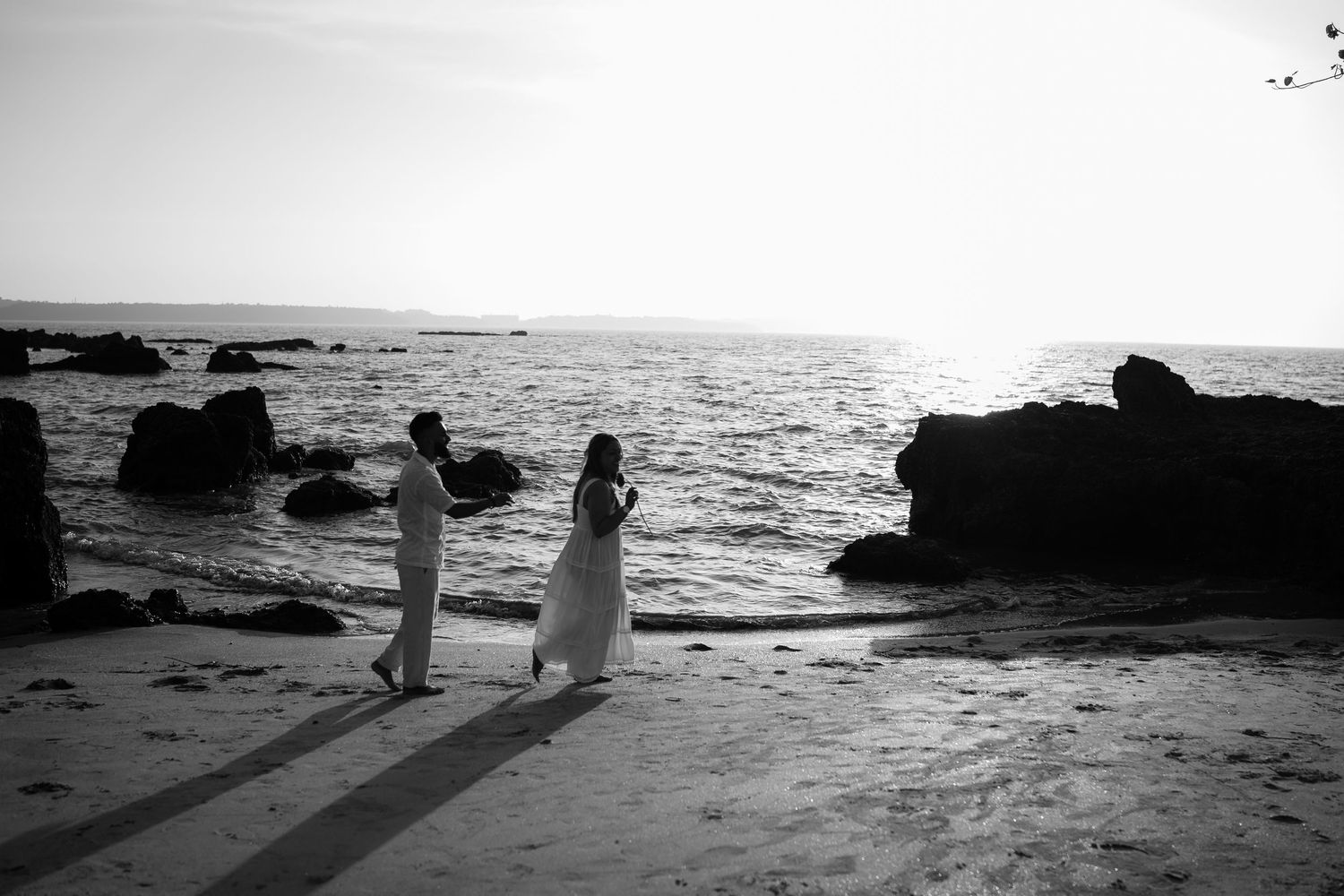 Two silhouetted figures walk along a rocky beach at sunset with ocean waves crashing in the background.