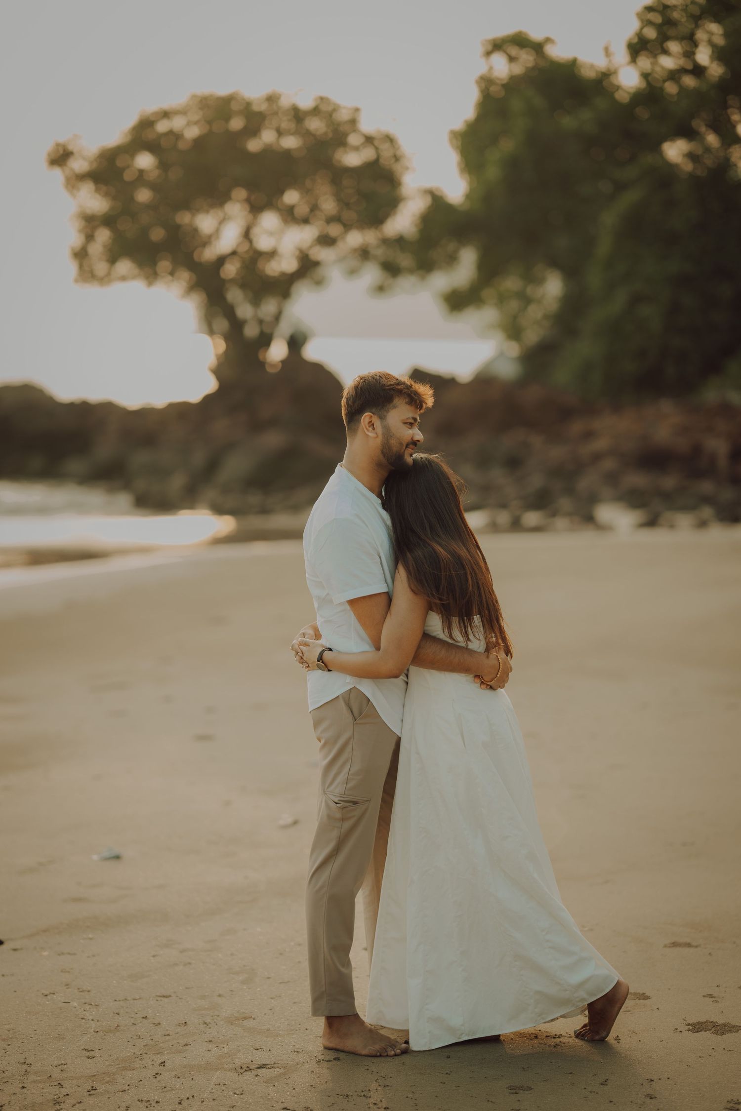 A couple shares an intimate moment on a sandy beach during golden hour as sunlight illuminates their silhouettes.