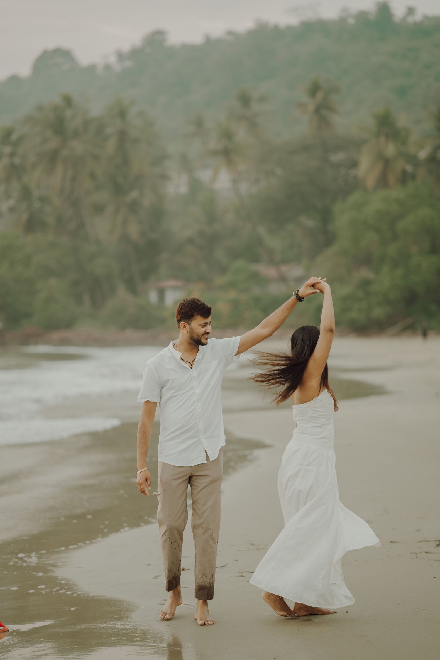 A couple in white attire walking and dancing together on a misty tropical beach with palm trees in the background.