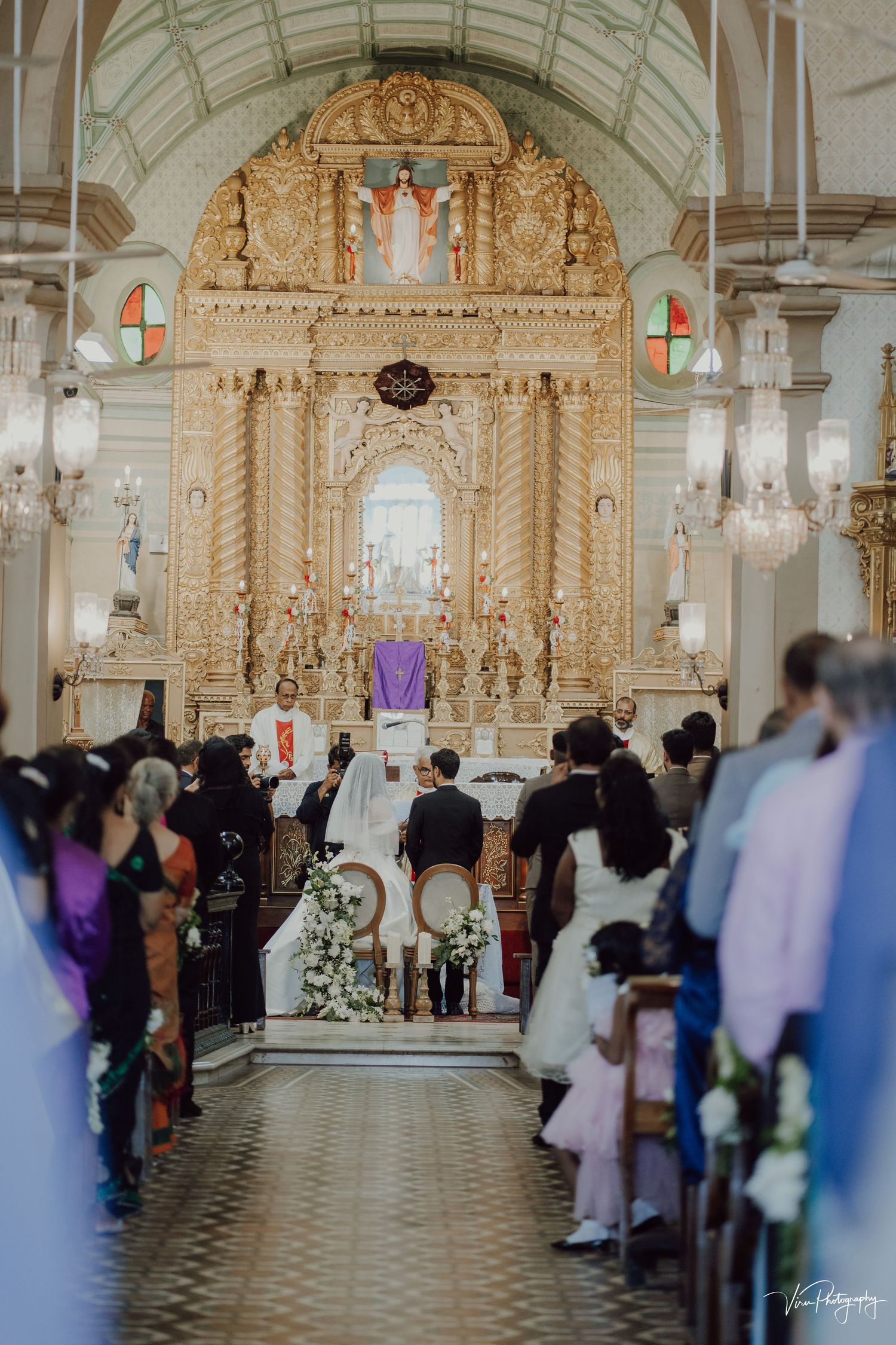 A church congregation gathers inside an ornate white Catholic church with baroque altar and decorative architecture during a service.