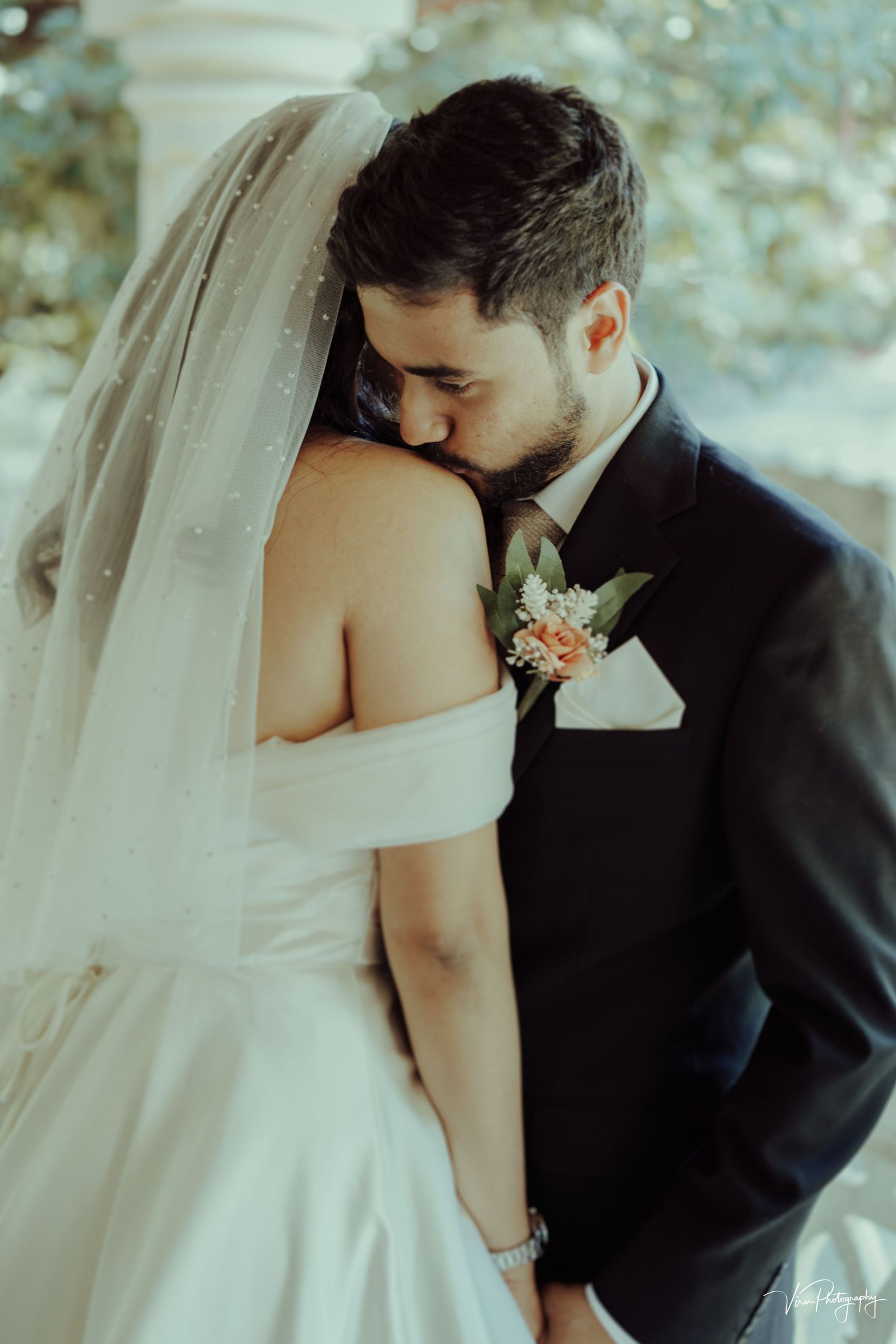 Two newlyweds share an intimate moment as the groom tenderly embraces his bride in her white off-shoulder gown and veil.