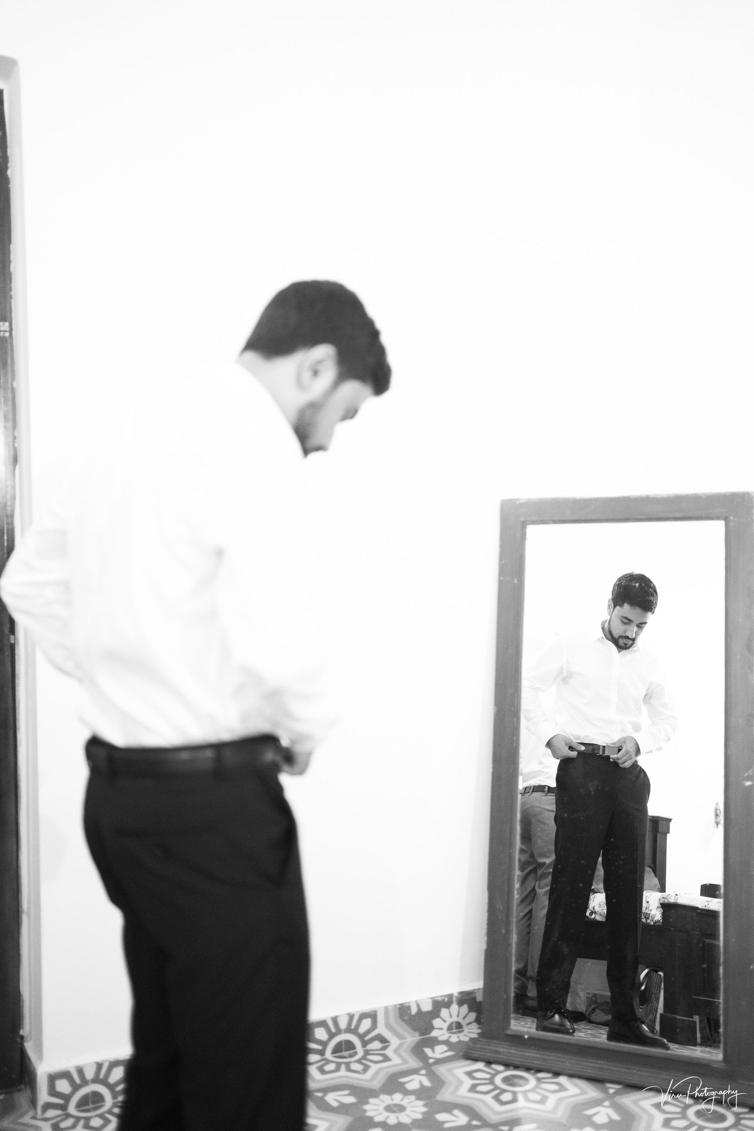 A black and white photograph of a person in formal attire looking down while standing next to a mirror reflection.