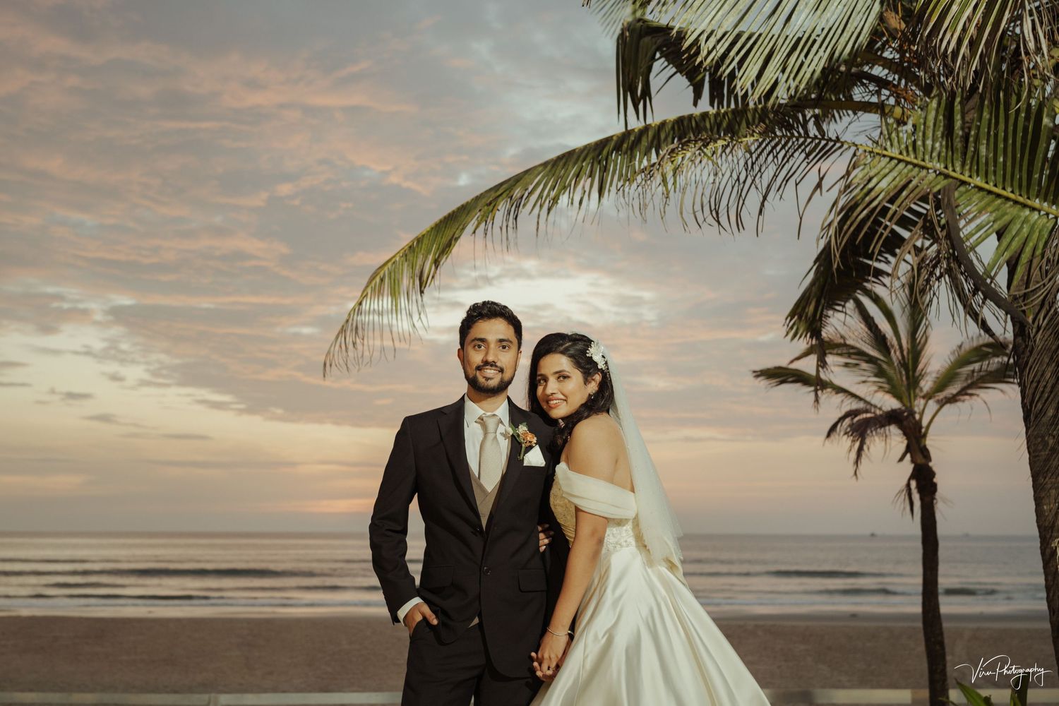 A newlywed couple stands on a beach at sunset with palm trees and the ocean in the background.