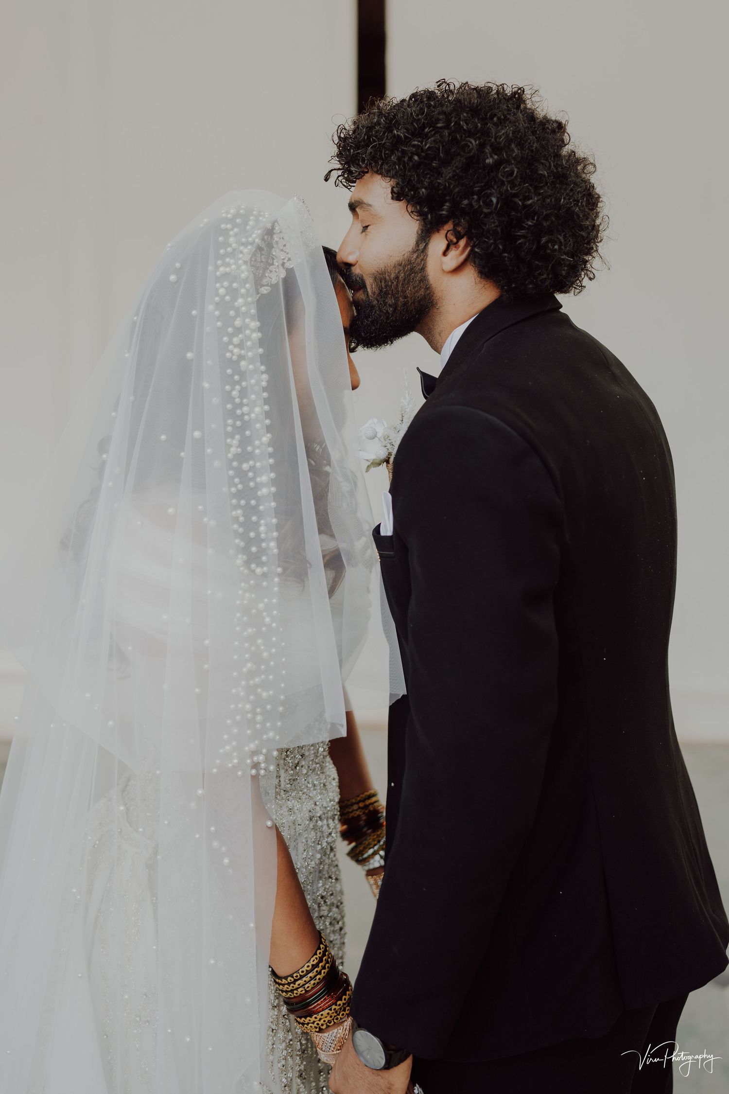 Tender moment captured at wedding as groom in black suit leans in to kiss bride wearing delicate beaded veil.