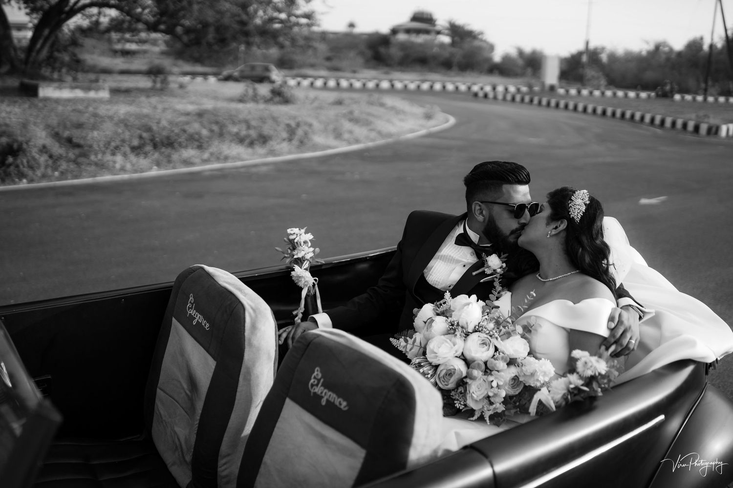 Newlywed couple sharing an intimate moment in a classic convertible car driving down a winding road in black and white.