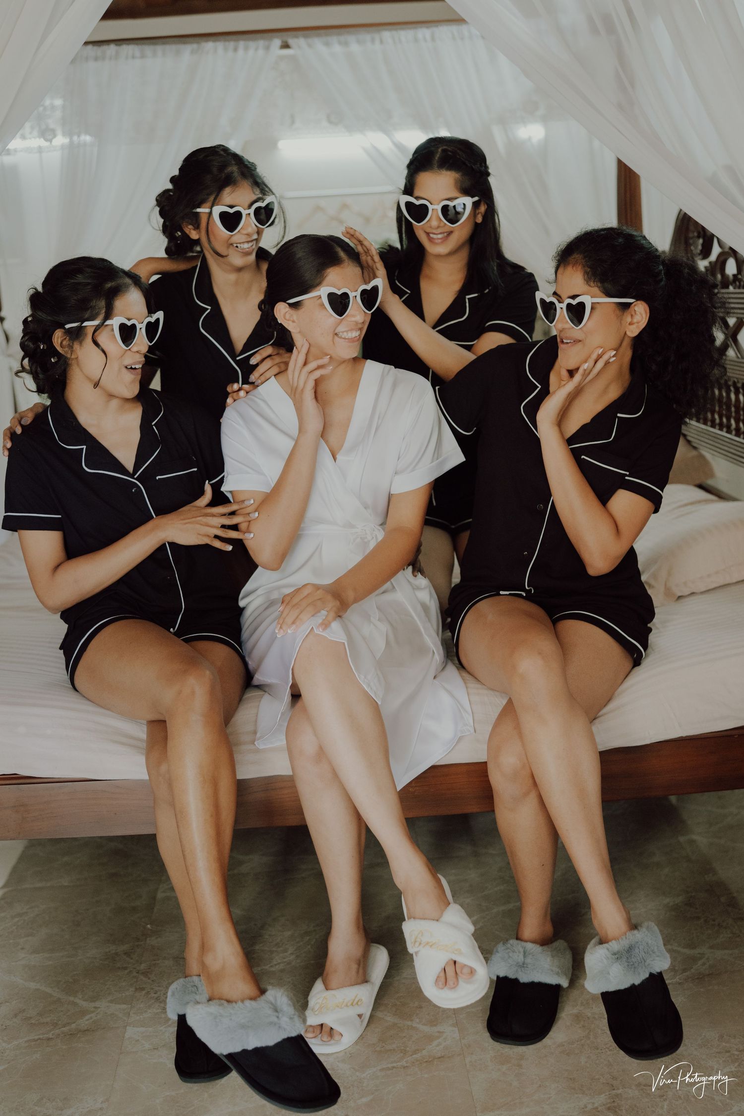 Group wearing matching heart-shaped sunglasses and cozy loungewear pose together on white furniture for fun bridal party photo.