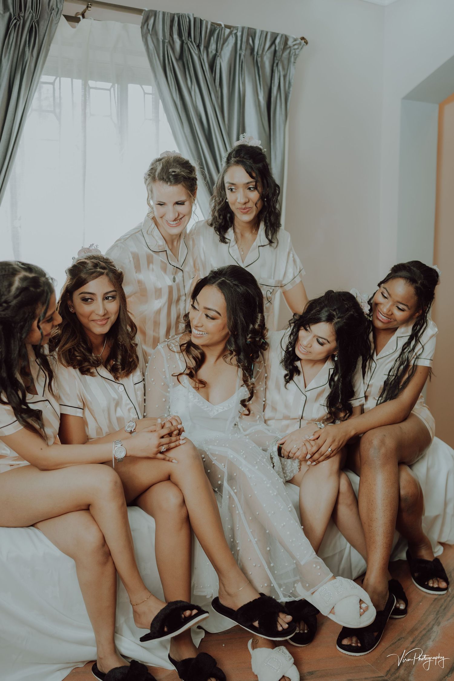 Group wearing satin robes gather together on bed during wedding day preparations in natural light.