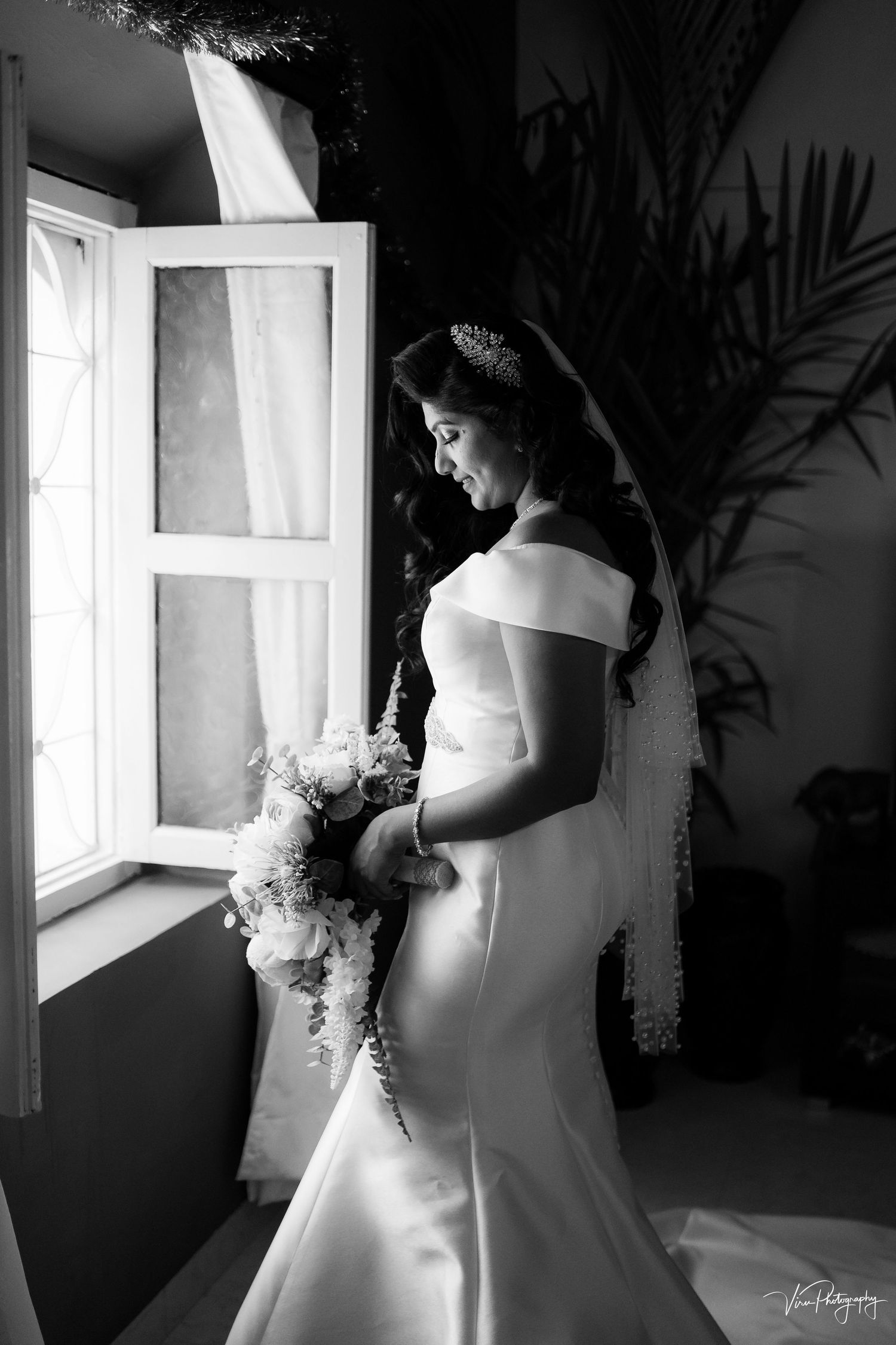 Elegant black and white portrait of bride in off-shoulder wedding gown standing by window with bouquet.