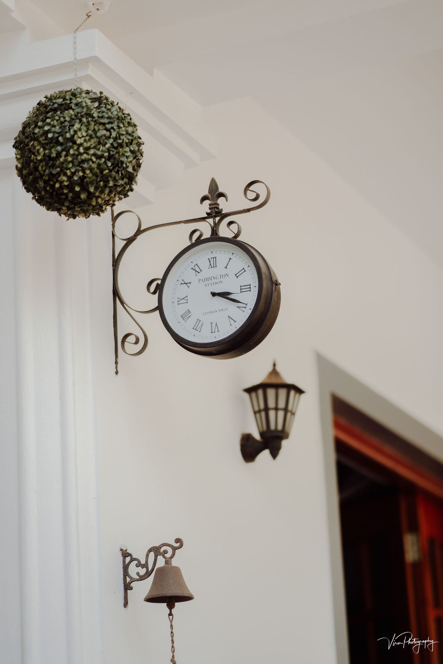 Vintage-style wall clock, lantern light, and decorative bell mounted on white wall with a green topiary ball above.