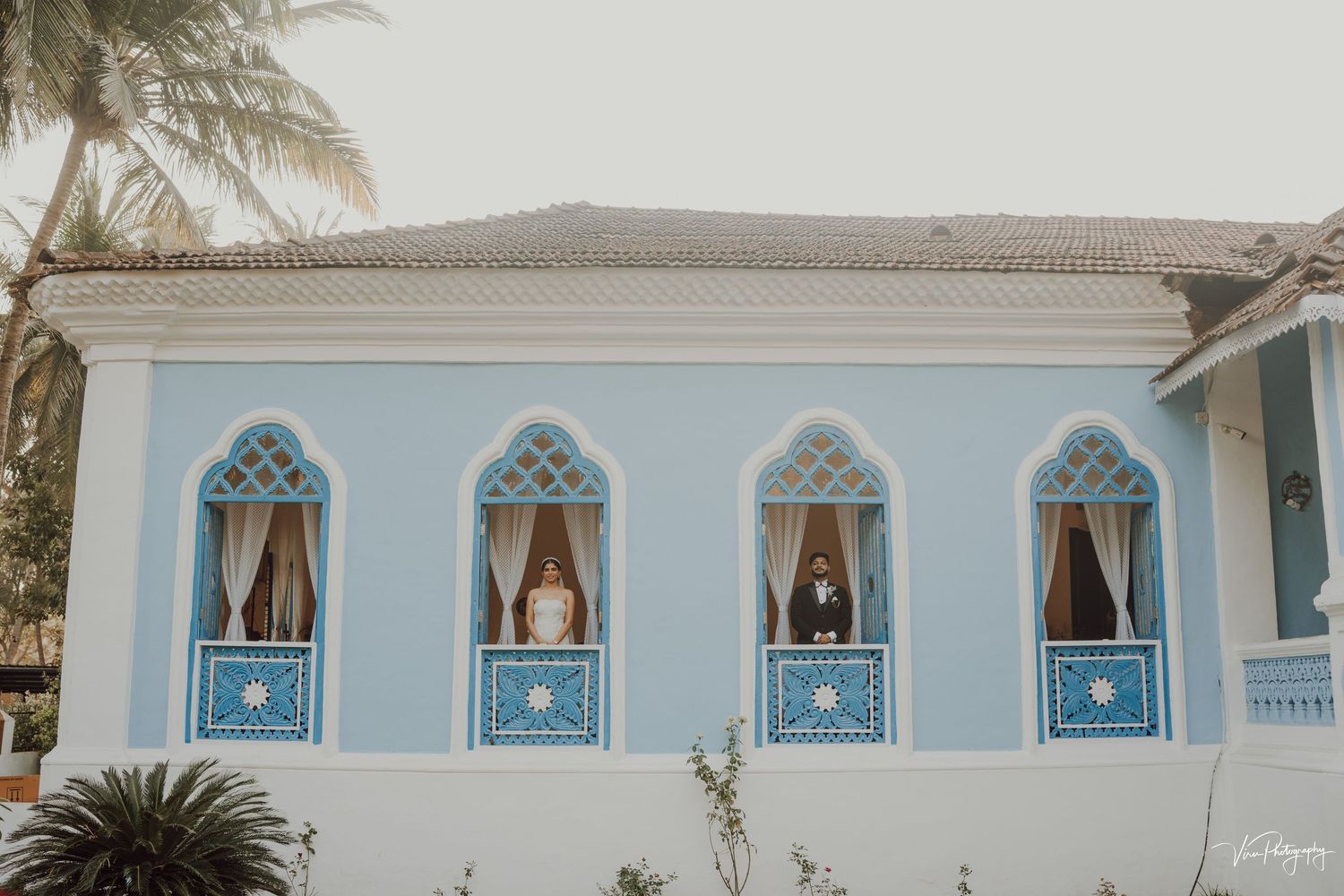 Light blue colonial style building facade with four ornate arched windows featuring decorative turquoise balconies and curtains.
