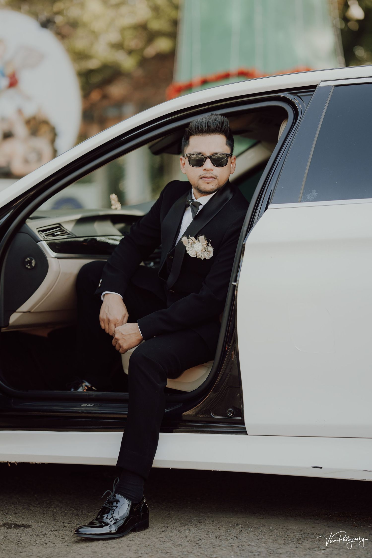 A stylishly dressed man in a black suit and sunglasses poses confidently from the driver's seat of a white luxury car.