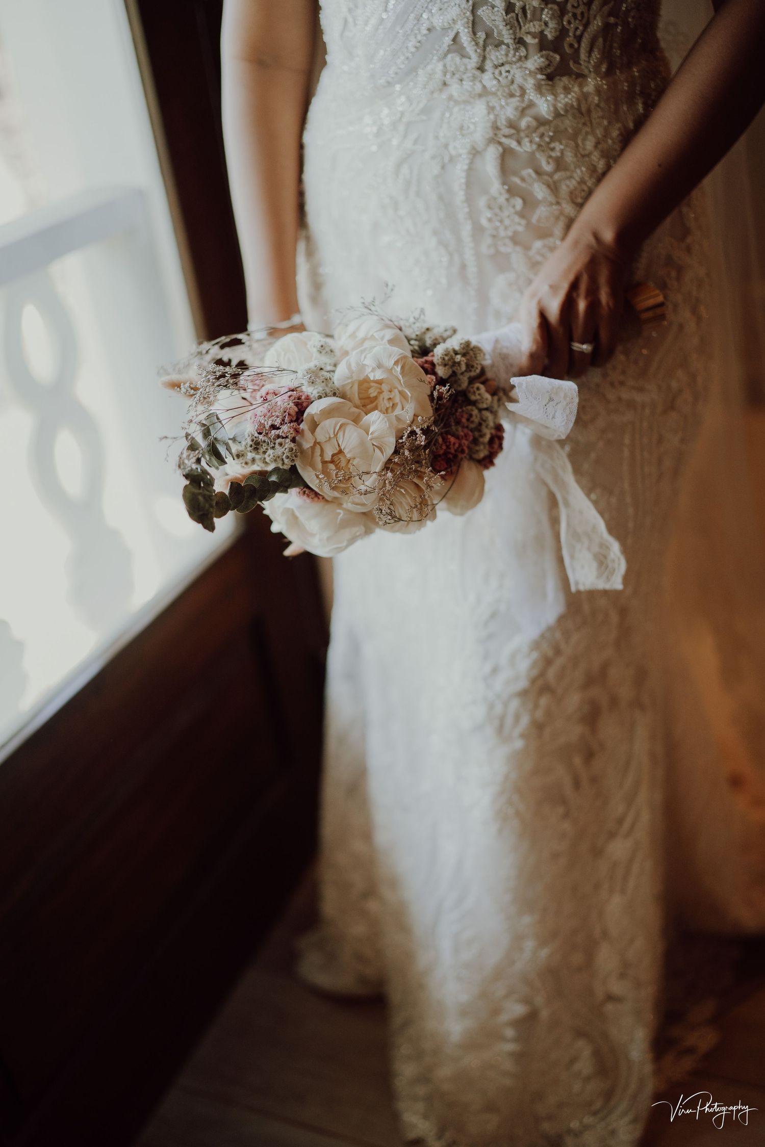 A bride in a lace wedding dress holds a delicate bouquet of soft pink and white flowers wrapped in ribbon.