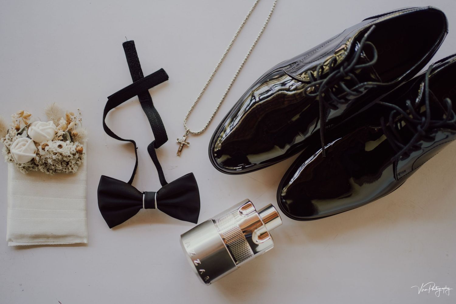 Elegant black dress shoes arranged with a bow tie, perfume bottle, and small white floral arrangement on a light surface.