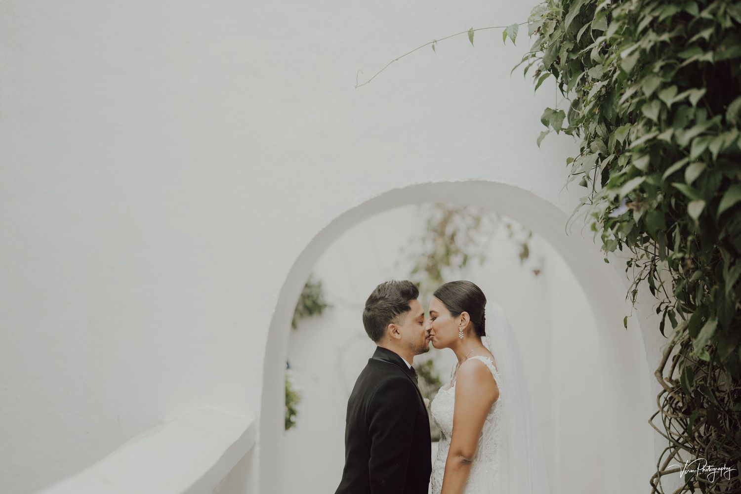 Two people share a tender moment under a white archway surrounded by lush greenery.