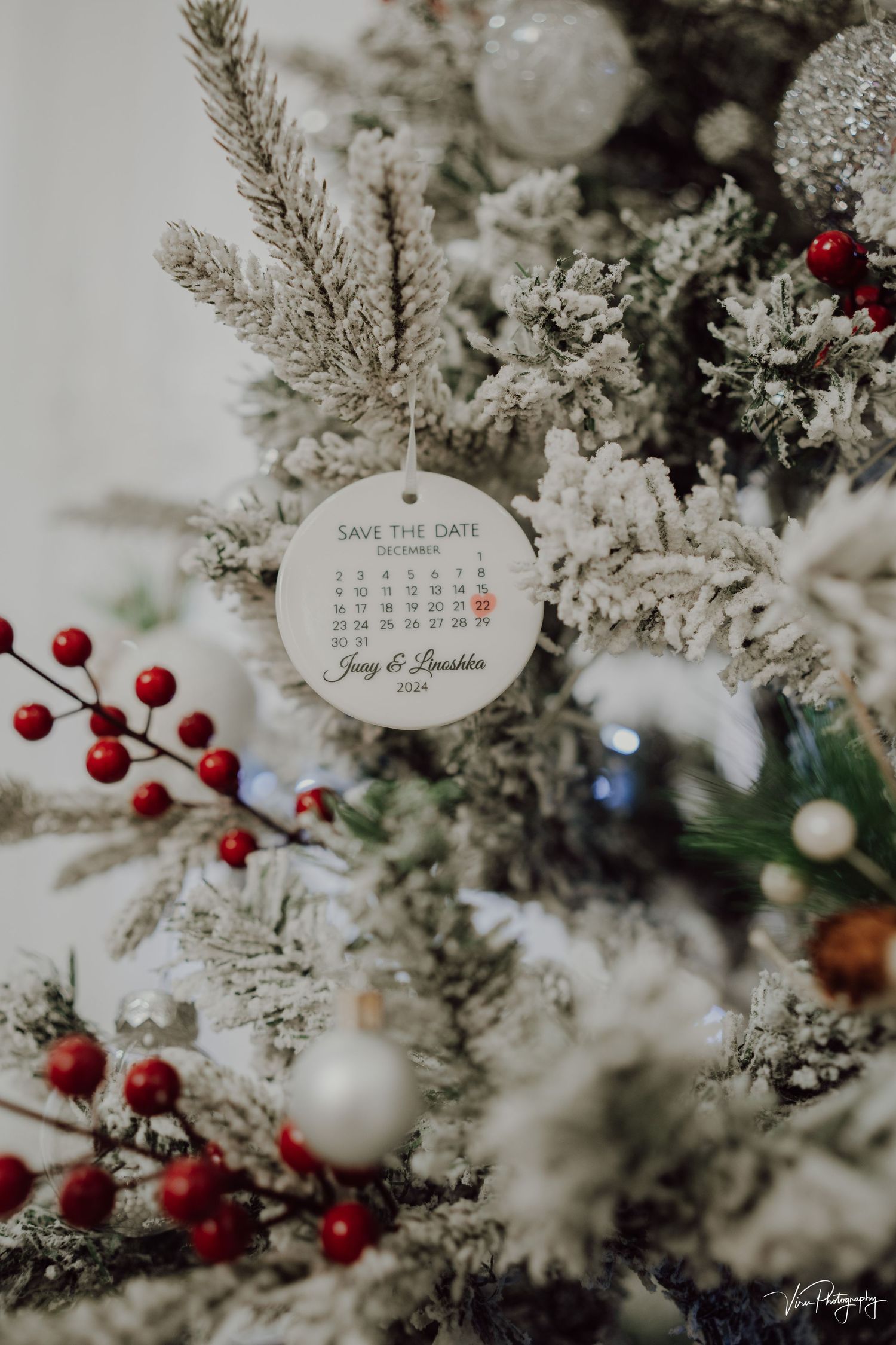 A save the date calendar ornament hangs on a frosted Christmas tree with red berries and snowy pine branches.