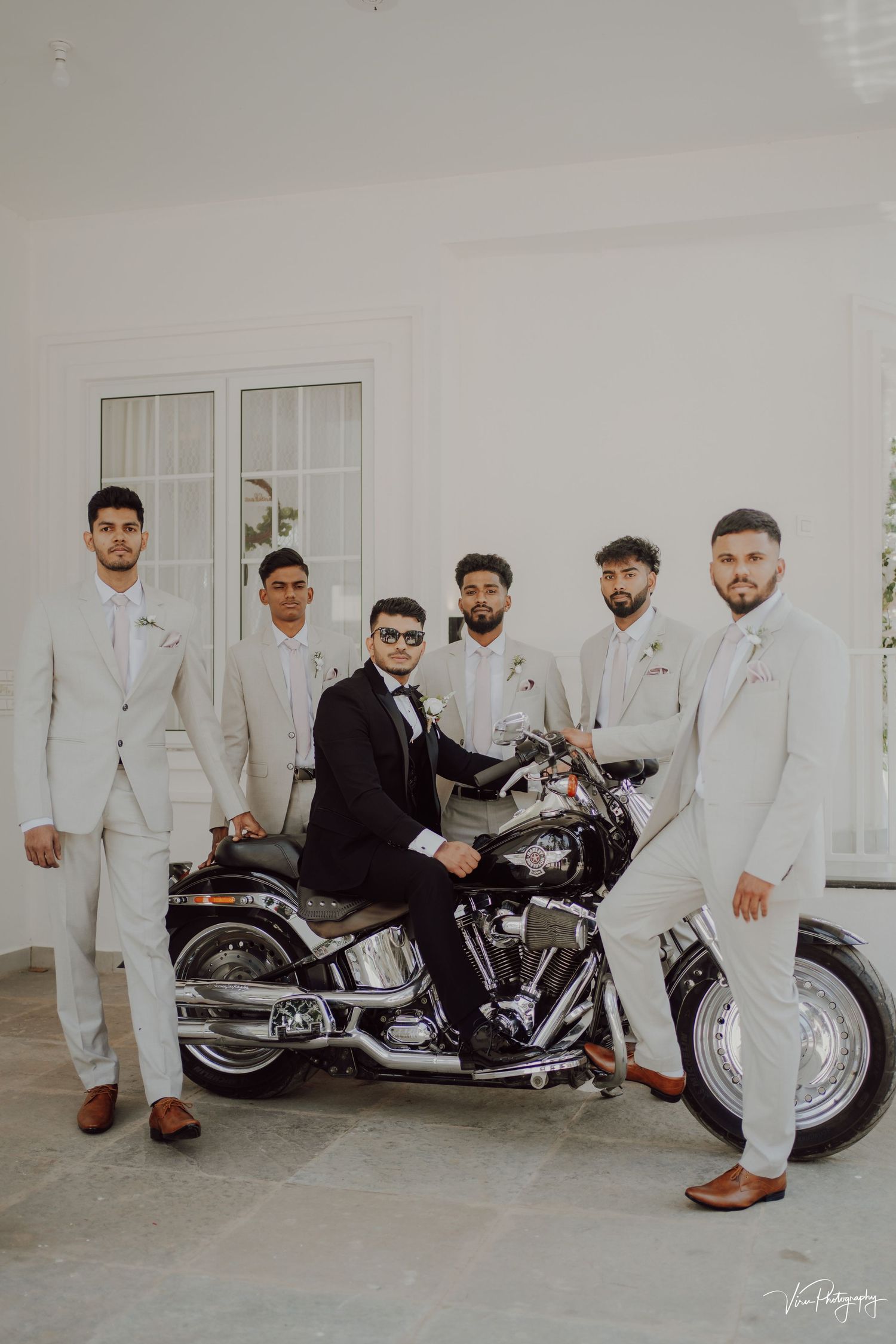 Groomsmen in white suits pose with one in black next to a classic motorcycle at an elegant venue.