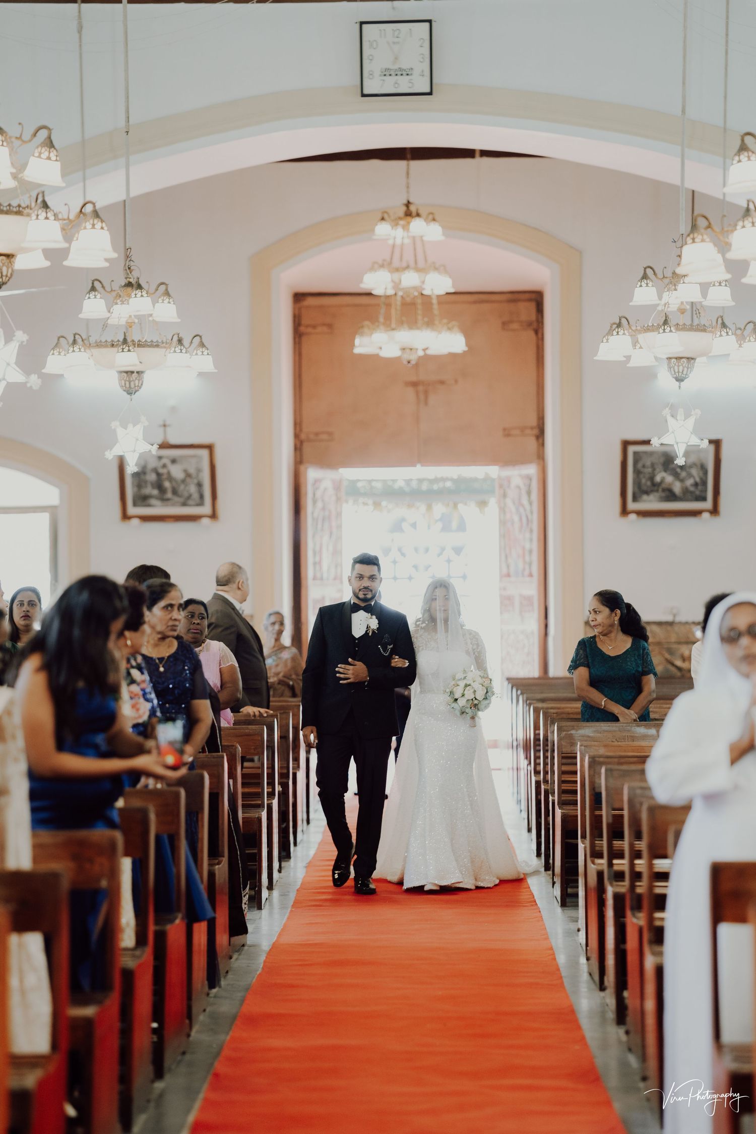 A traditional church wedding ceremony with a red carpet aisle, arched ceiling, and elegant chandelier lighting.