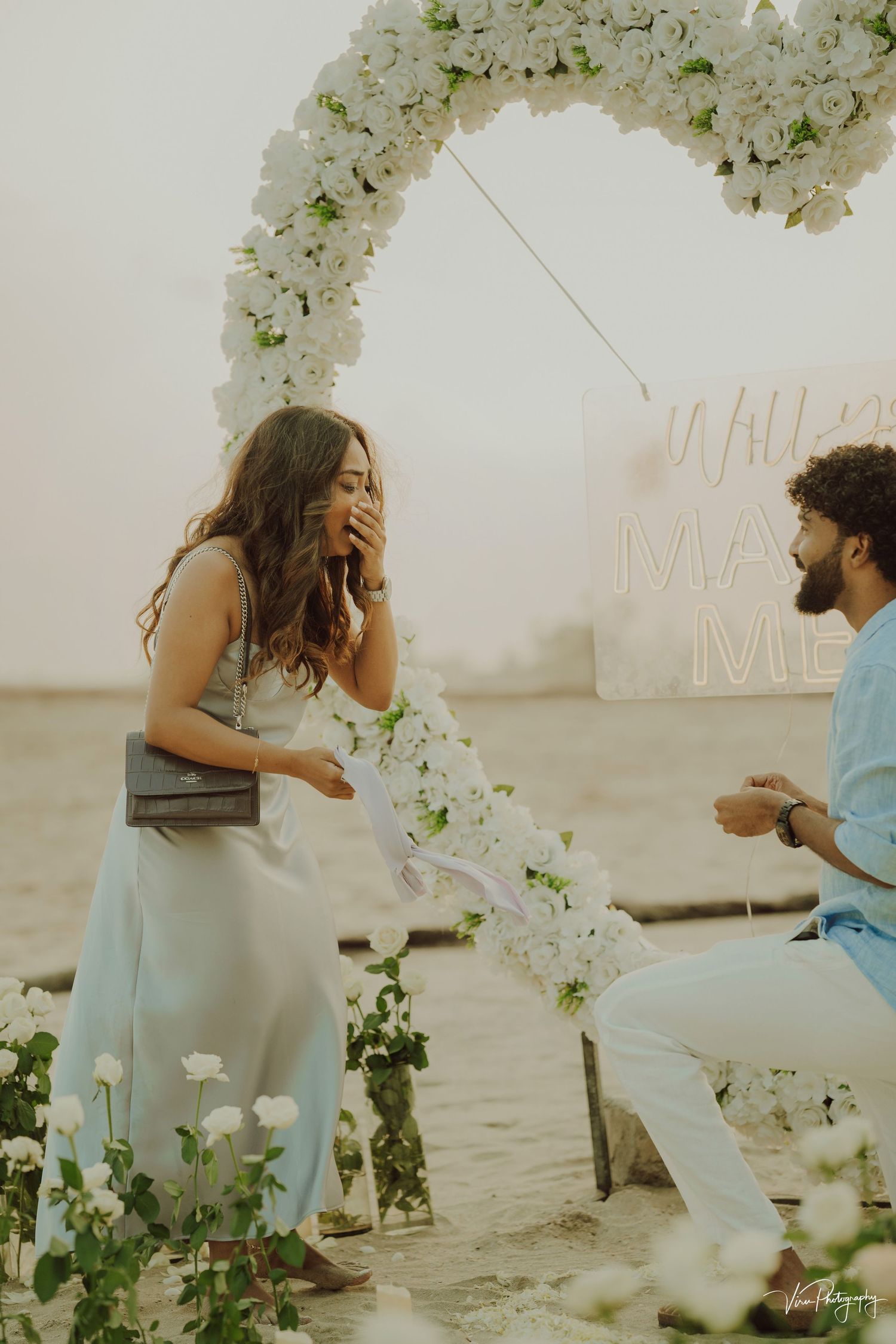 A romantic beach proposal scene with white floral arch decorations and sunset lighting.
