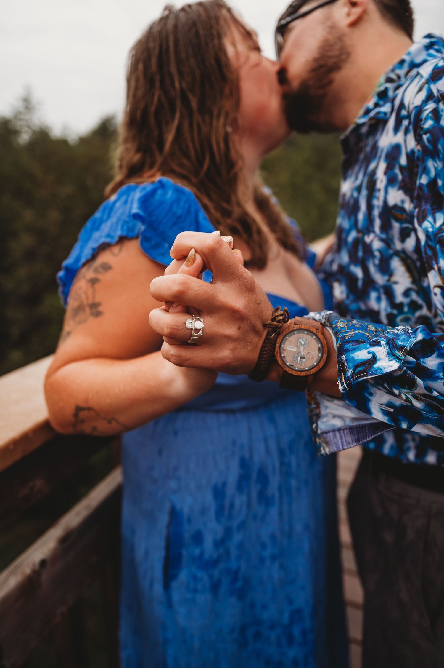 Couple holding hands in romantic moment on bridge, with blue dress and woodwatch detail visible in sunlight.