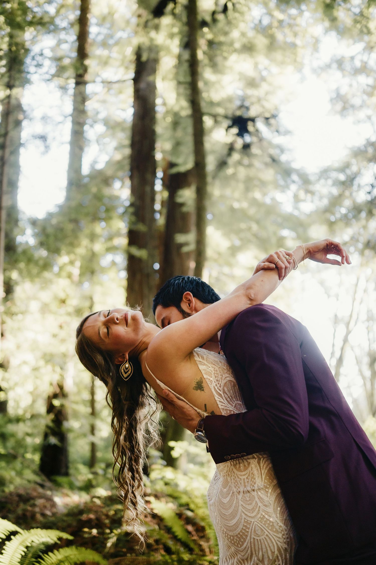 A romantic couple in formal attire share an intimate dance pose among towering trees in a sunlit forest setting.
