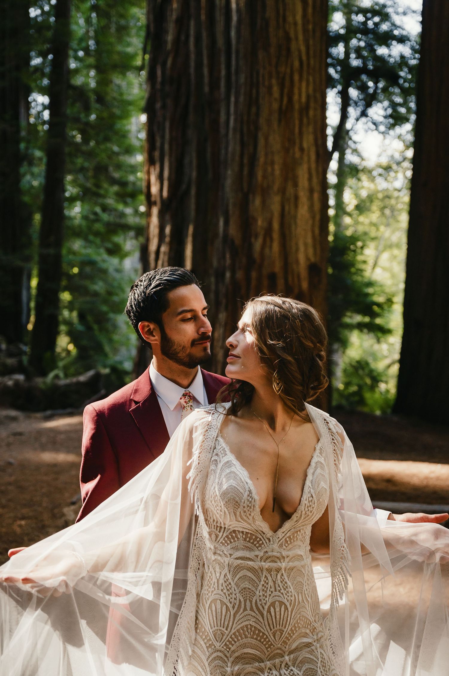 A romantic wedding couple shares tender moments among towering redwood trees in a sunlit forest setting.
