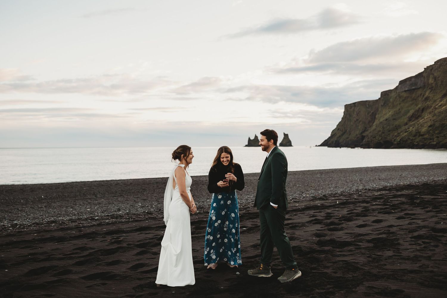 Silhouetted figures walk together on a dramatic black sand beach at sunset with rocky cliffs in the background.