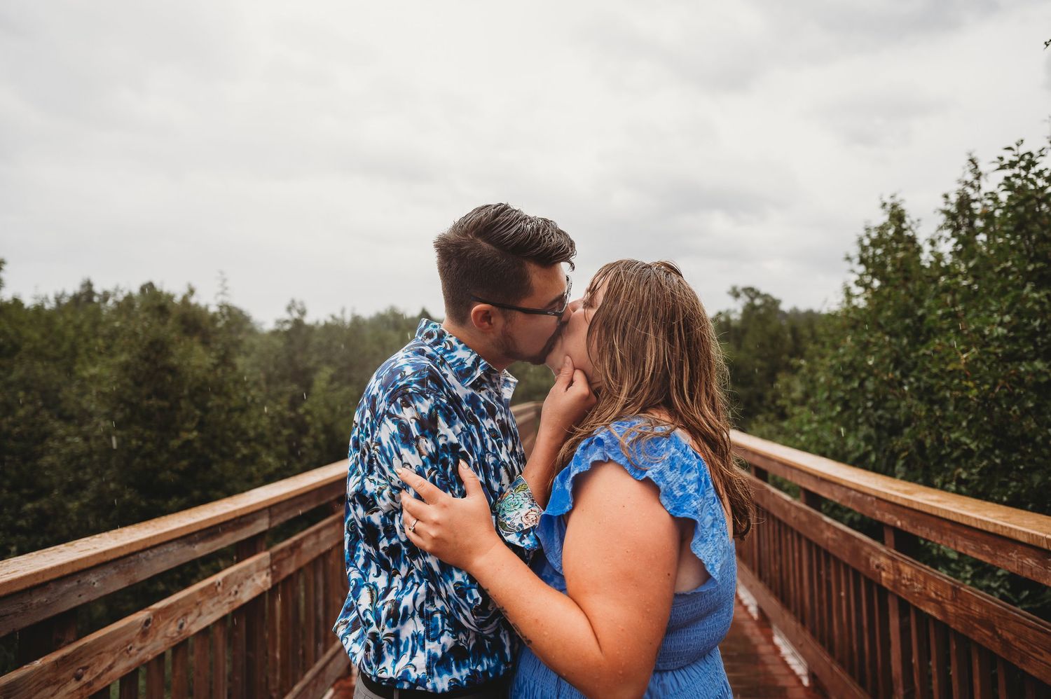 A romantic kiss takes place on a wooden bridge overlooking a forested landscape on a cloudy day.