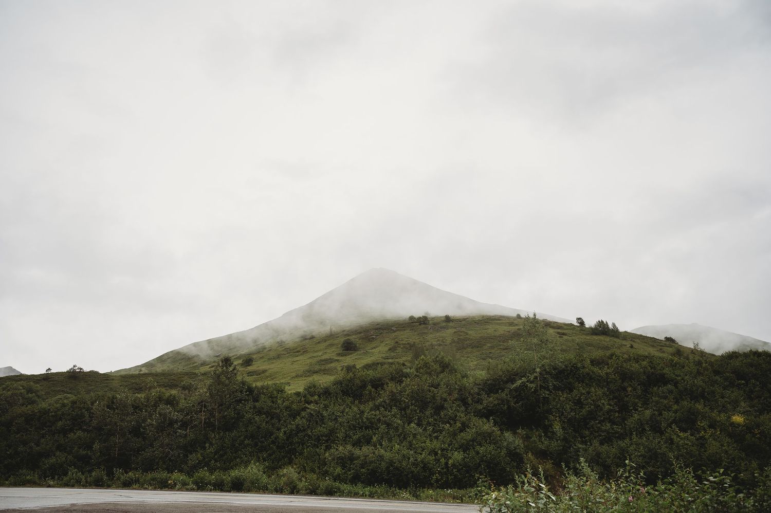 Misty mountain peak rises above green rolling hills with cloudy sky in the background.