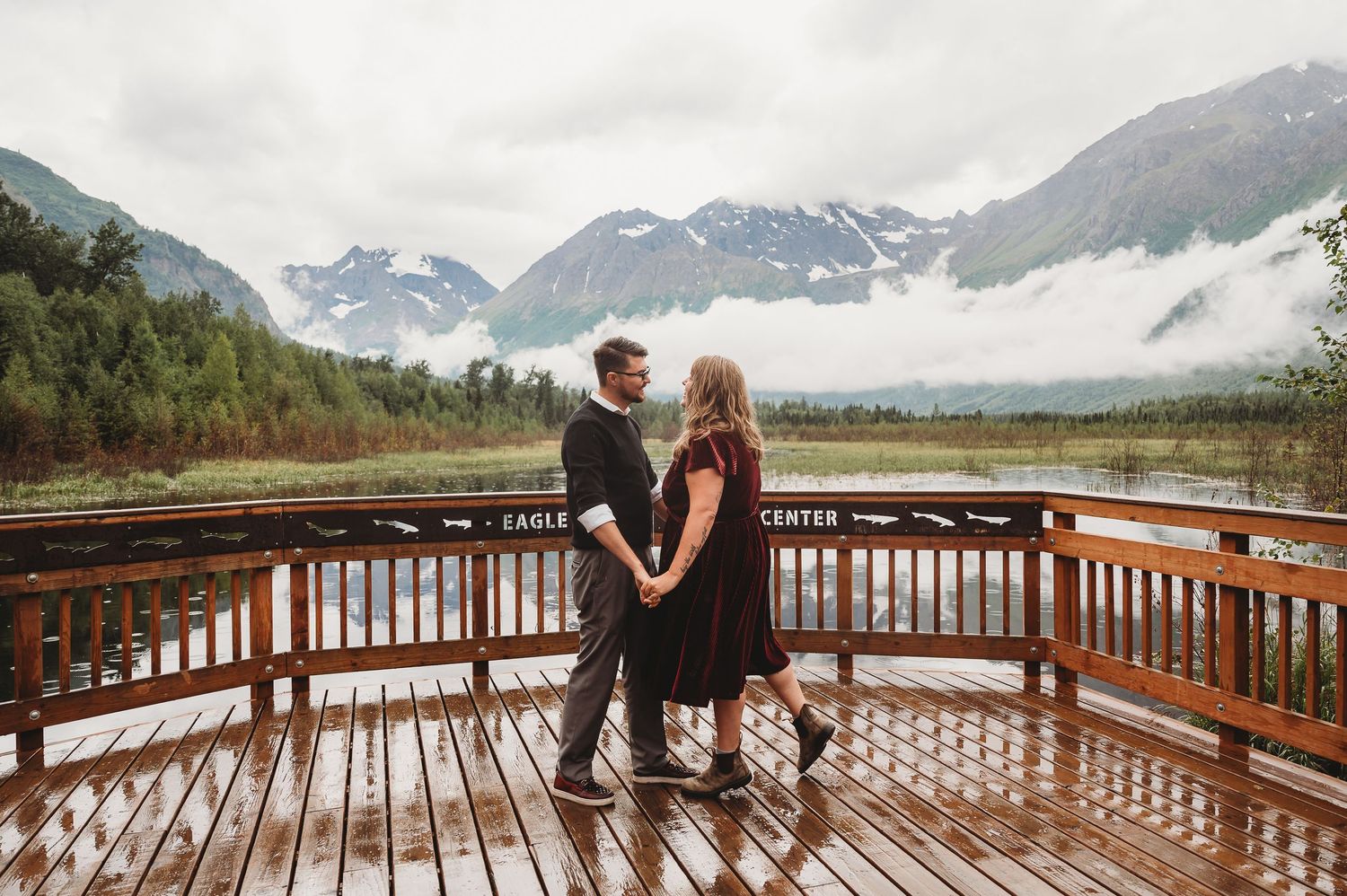 Couple stands together on wet wooden deck with snow-capped Alaskan mountains and low clouds in background.