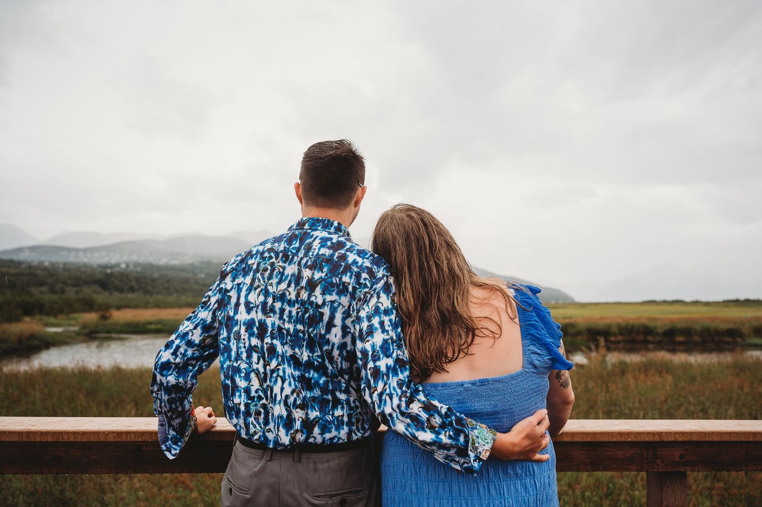 A couple in matching blue outfits embraces while looking out over a scenic mountain landscape on a cloudy day.