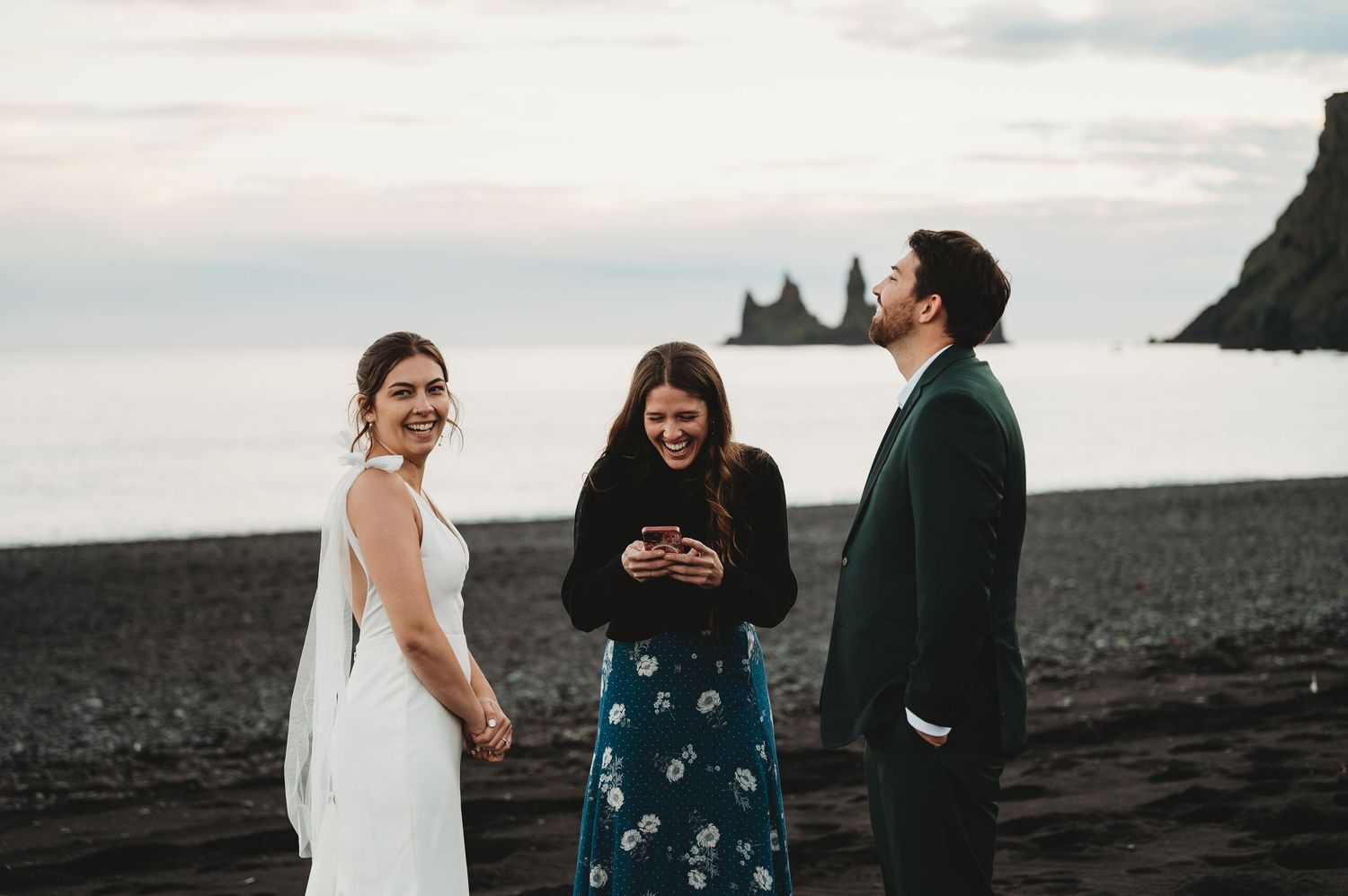 Laughing people gathered on a scenic beach in wedding attire during a celebration by the ocean.