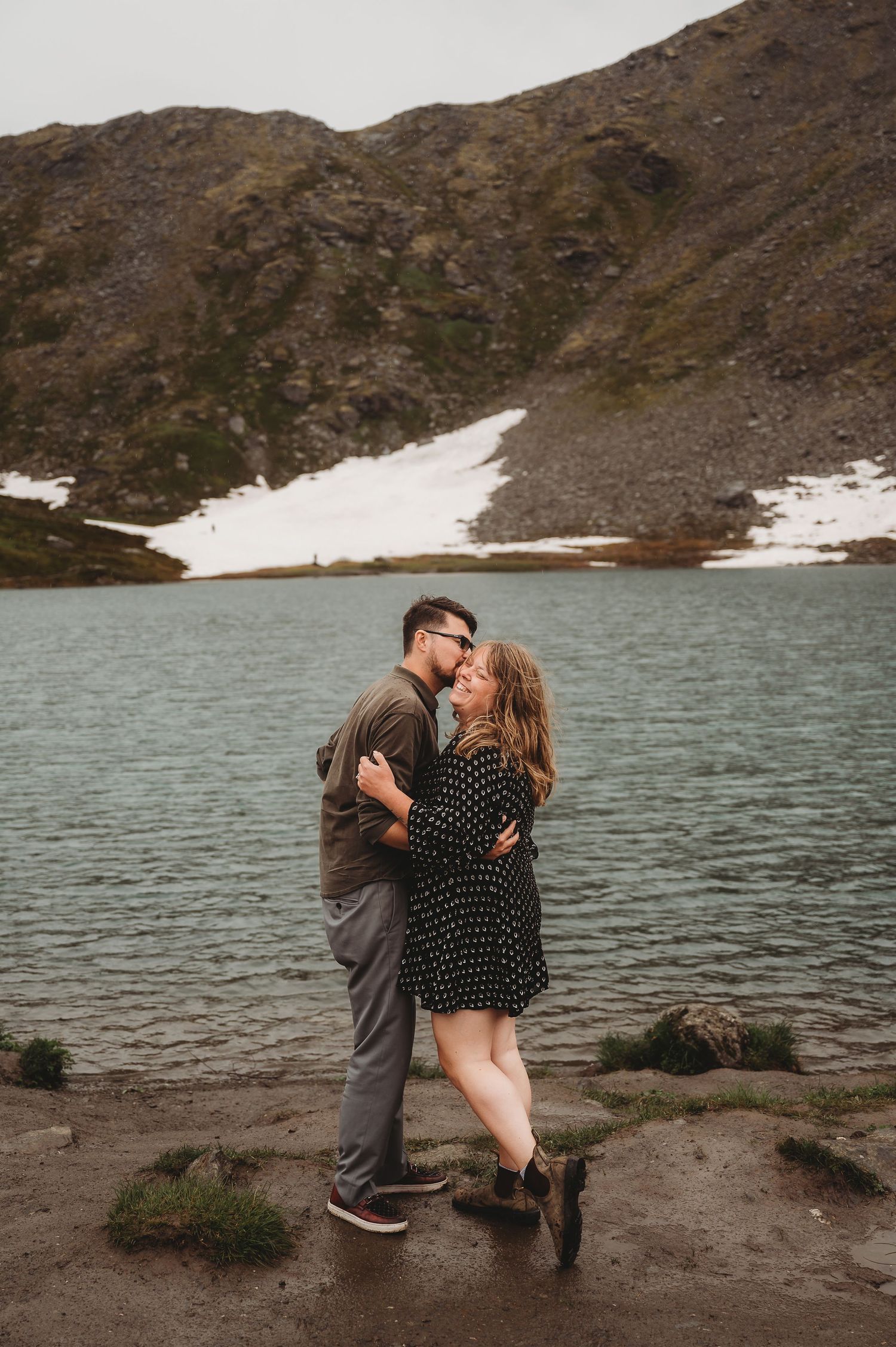 A couple shares a romantic embrace on rocks by an alpine lake with snowy mountains in the background.