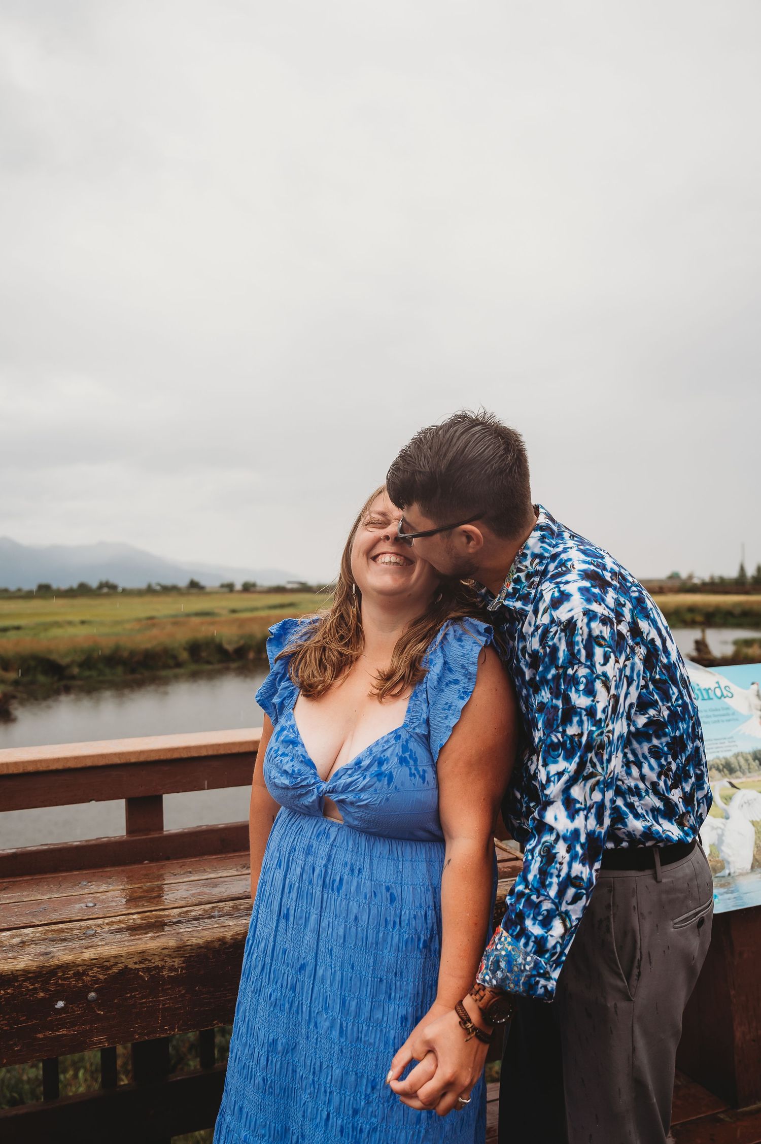 A couple shares an intimate moment on a wooden deck overlooking marshland and mountains on a cloudy day.