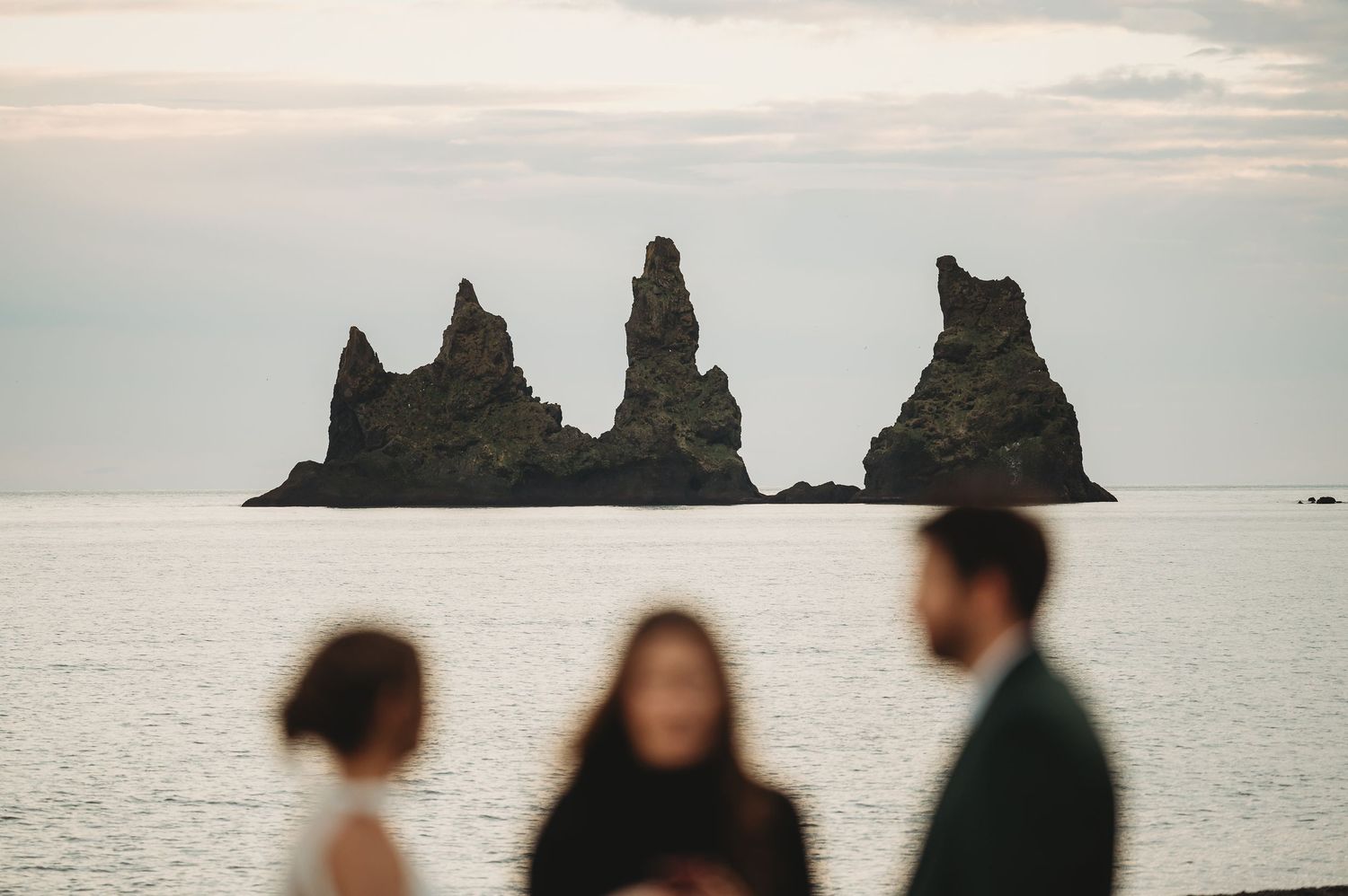 Silhouetted figures gather near ocean with iconic Reynisdrangar rock formations in background at dusk.