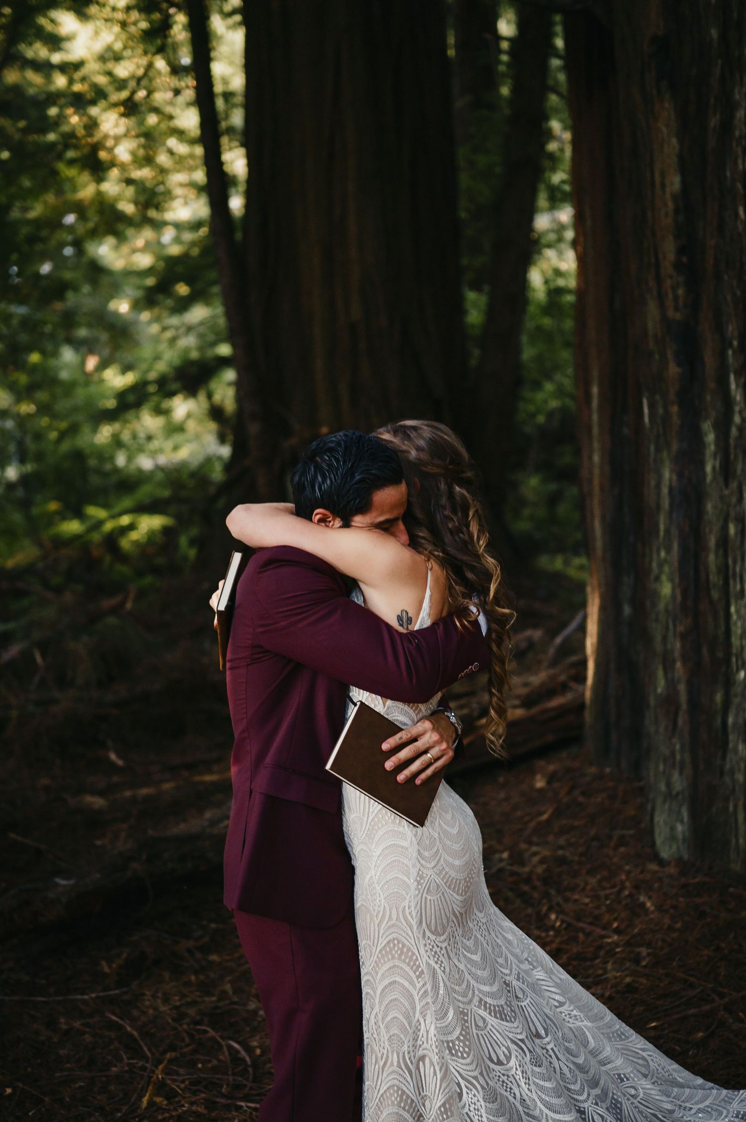 A romantic forest wedding moment captured in dappled sunlight as two figures embrace amongst towering redwood trees.