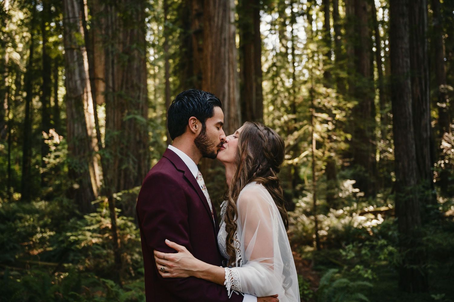A romantic wedding moment captured between a couple among majestic redwood trees in a sunlit forest.