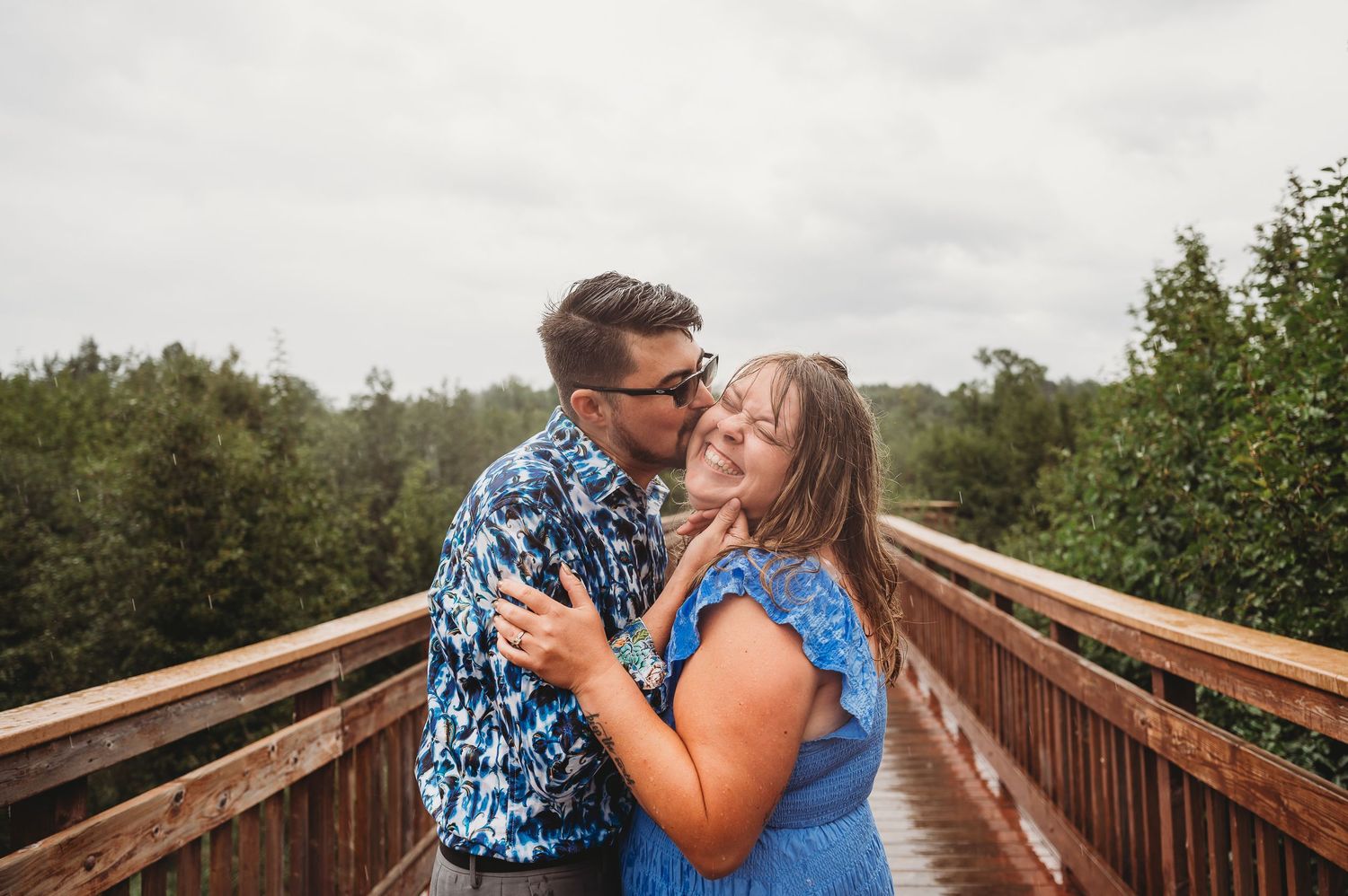 Couple wearing matching blue patterns share an intimate moment on a wooden boardwalk surrounded by green foliage.