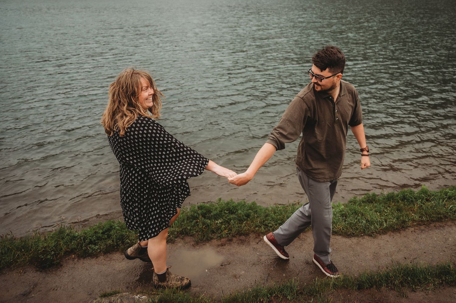 A couple holds hands while walking along a lakeside path in casual attire on an overcast day.