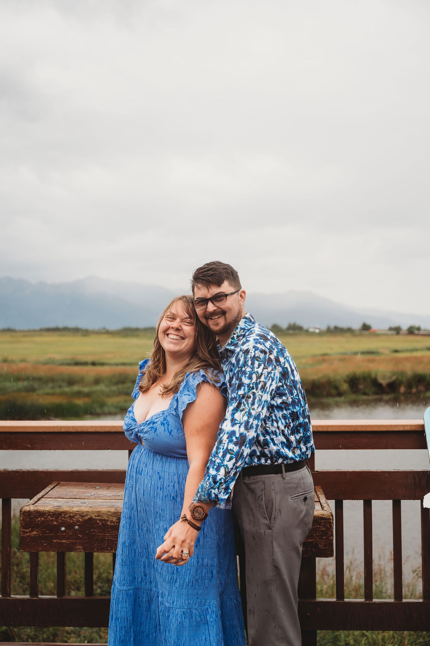 A couple in coordinated blue outfits share a tender moment while posing together on a wooden deck overlooking wetlands.