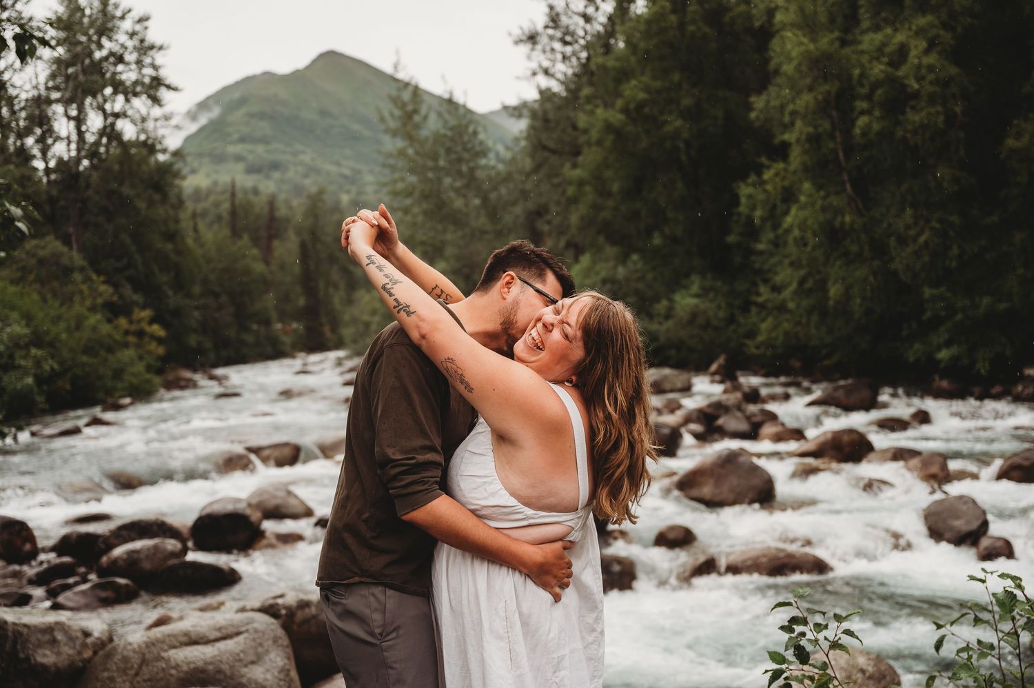 Two people embrace on a rocky riverbank in front of misty mountains during a romantic outdoor photo session.