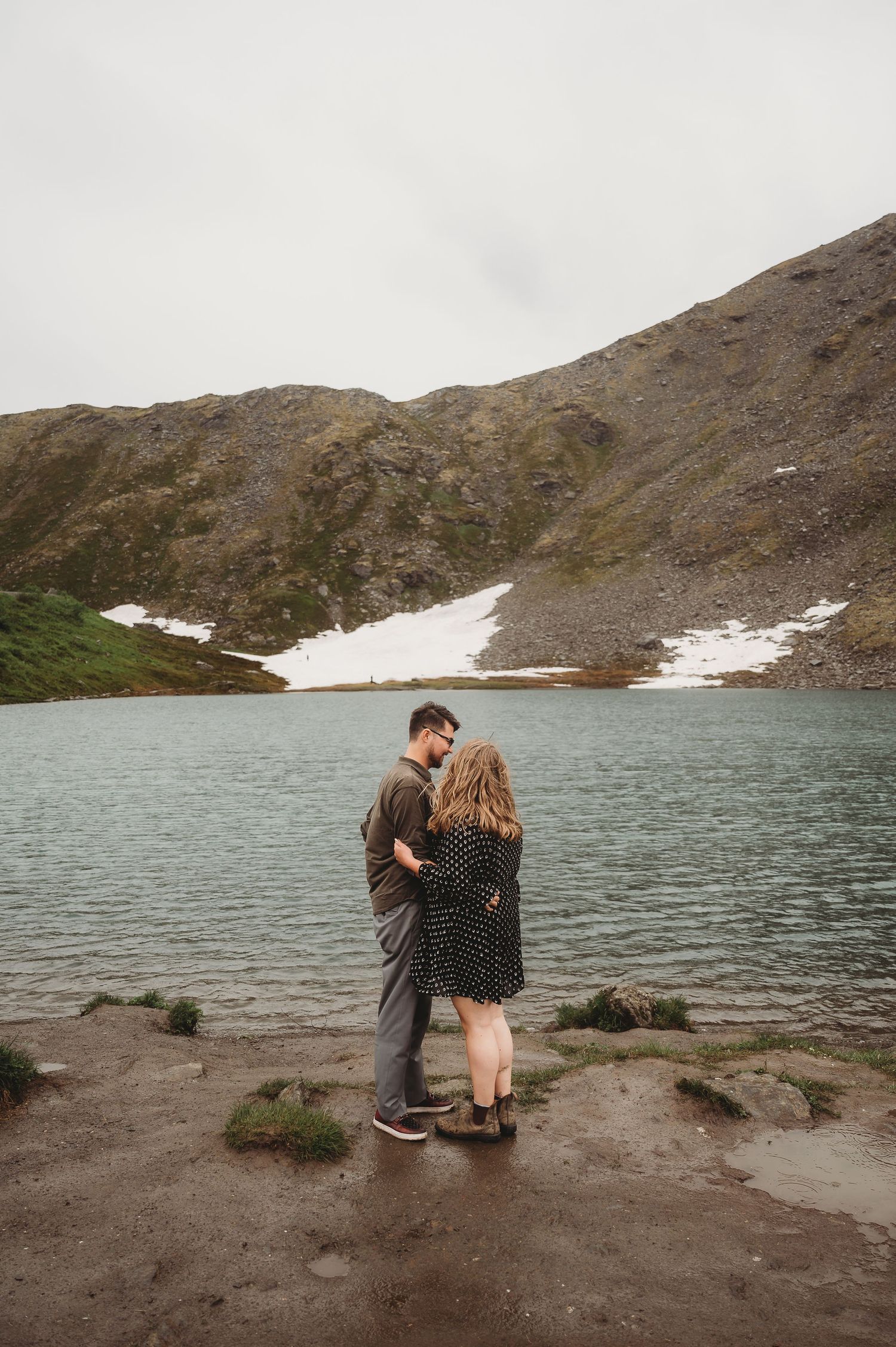 A romantic moment unfolds by an alpine lake surrounded by snow-capped mountains and rocky terrain.
