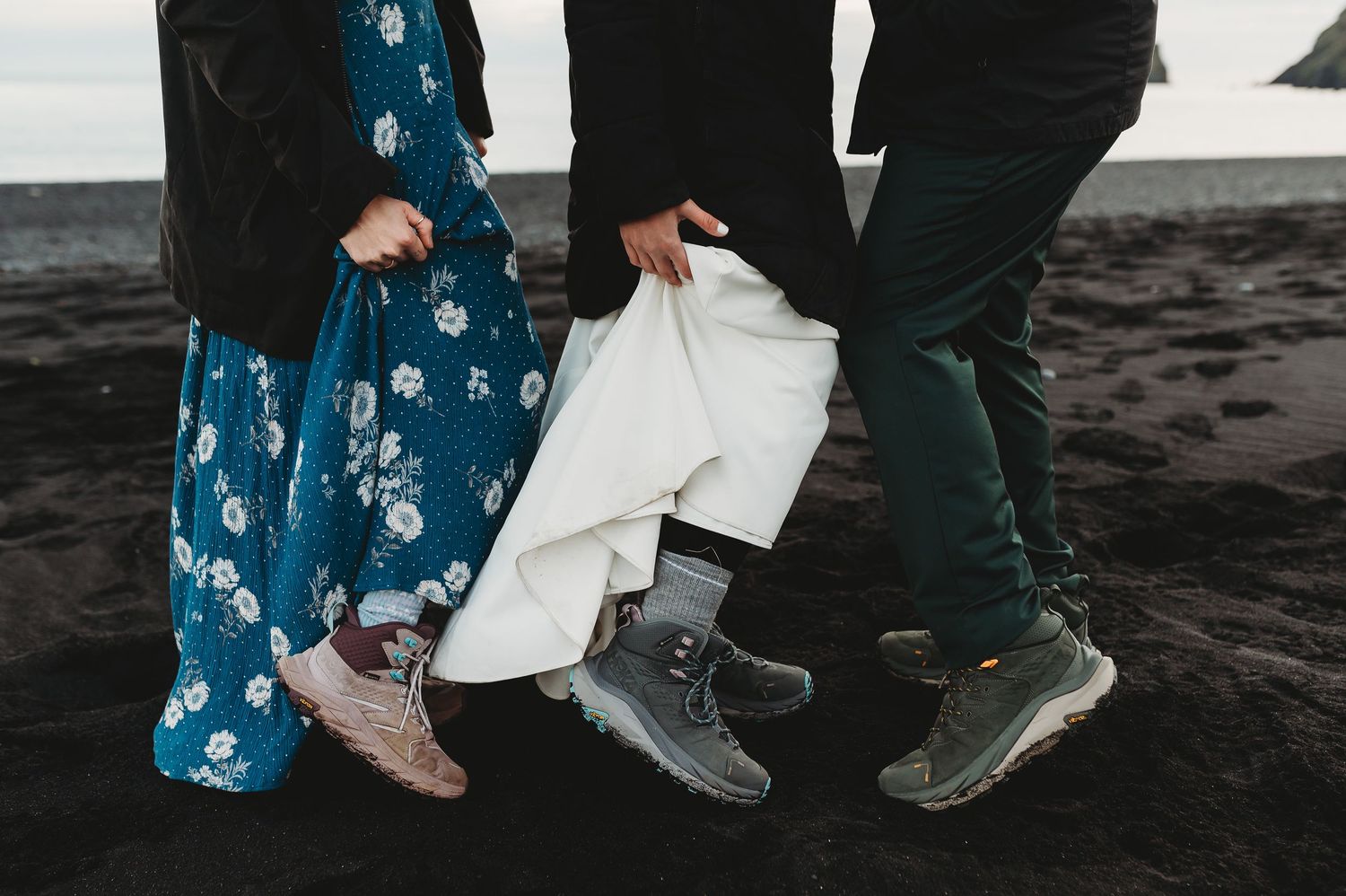 Two people in boots stand together on a black sand beach wearing floral blue dress and dark pants.