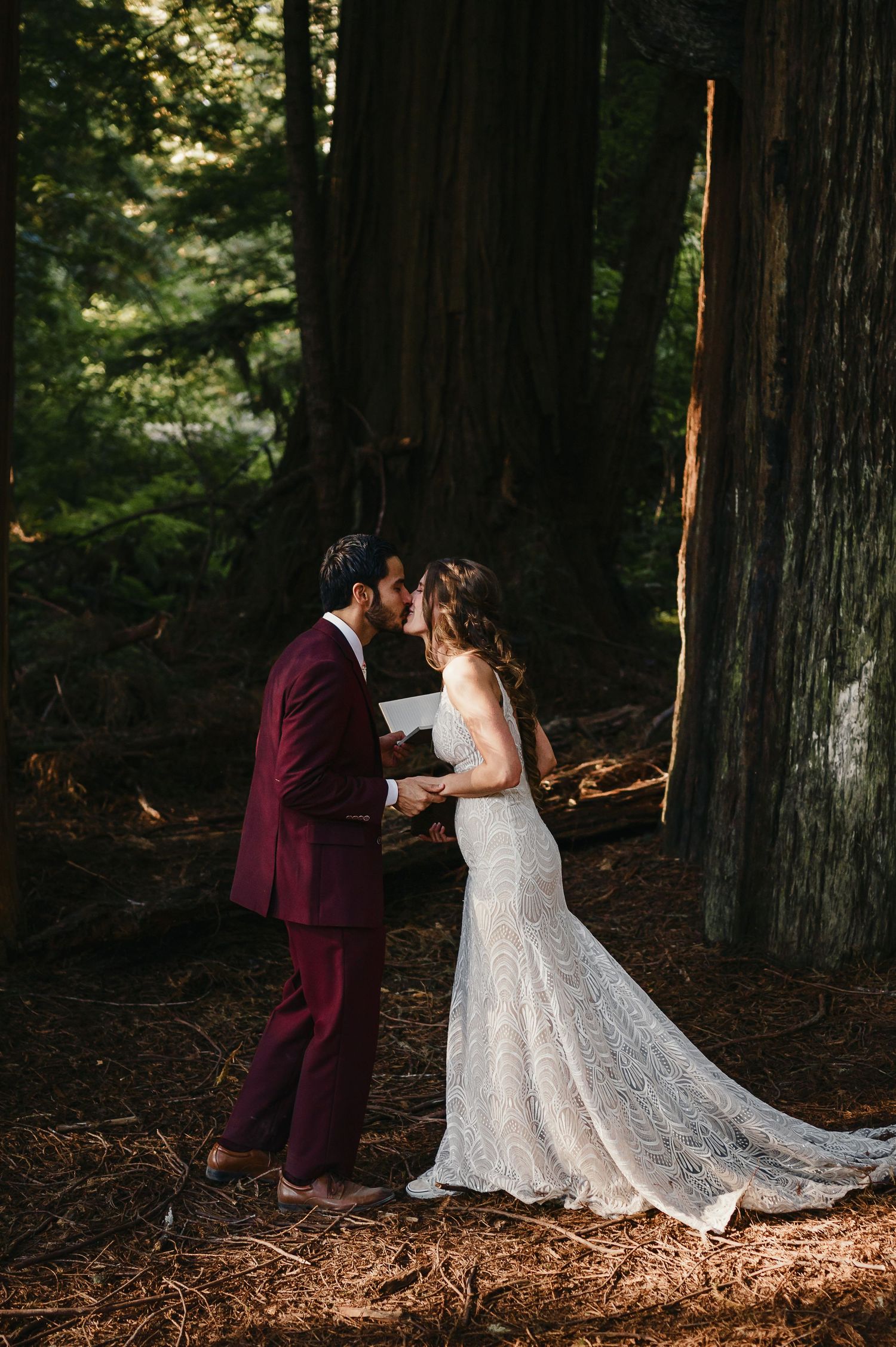 A couple shares an intimate moment in their wedding attire among towering redwood trees in a sunlit forest.