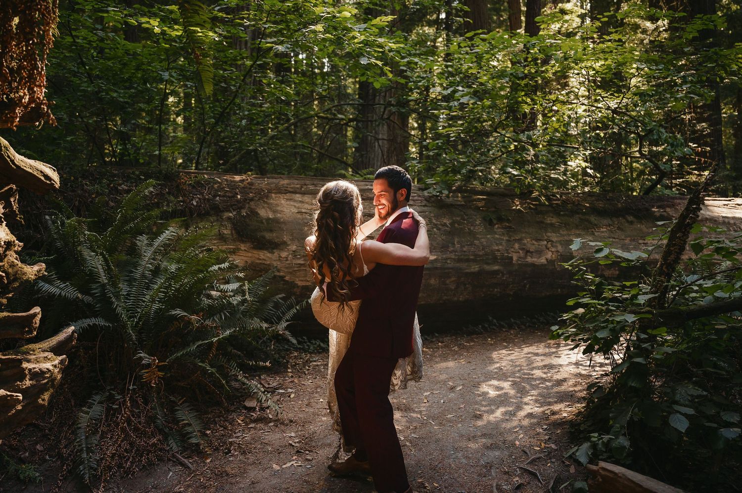 A person aims a compound bow in a wooded area with dappled sunlight filtering through the trees.