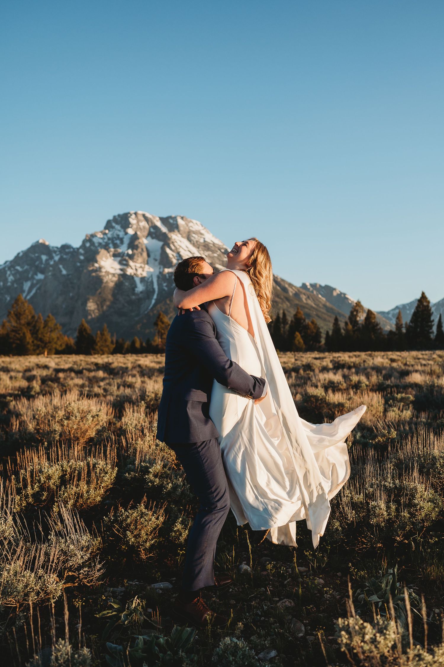 Groom lifts bride in white gown in a mountain meadow with snow-capped Grand Teton peaks under a clear blue sky.