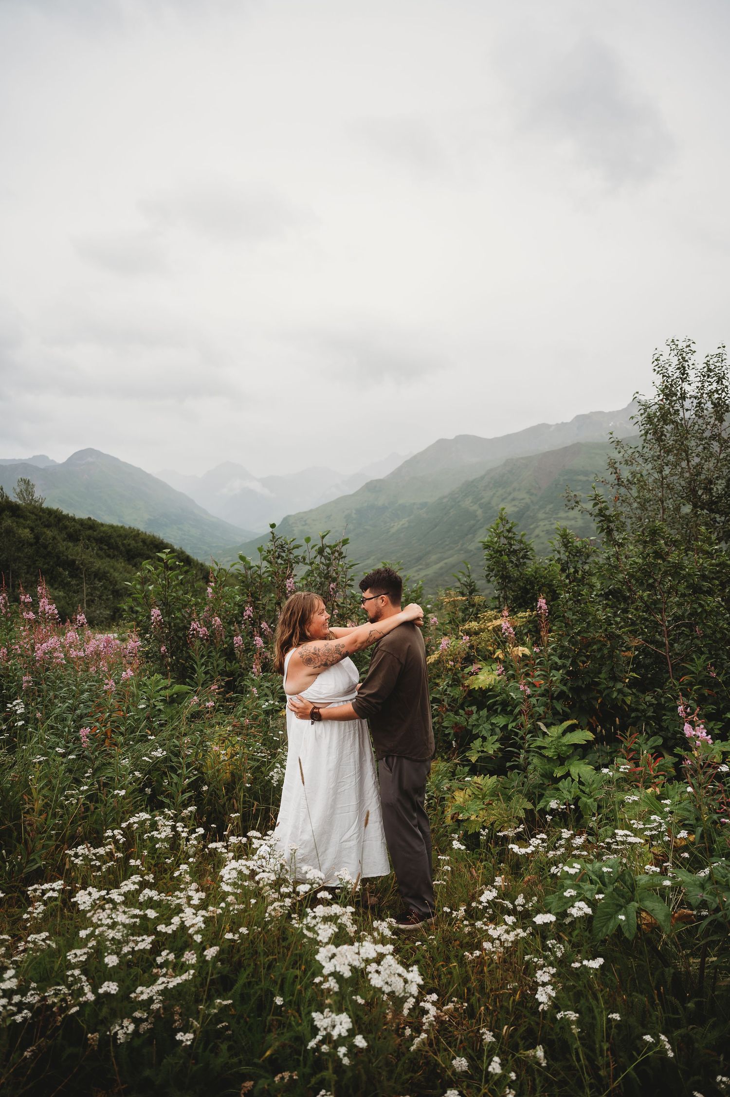 A couple embraces in a scenic mountain meadow filled with white wildflowers and green foliage.
