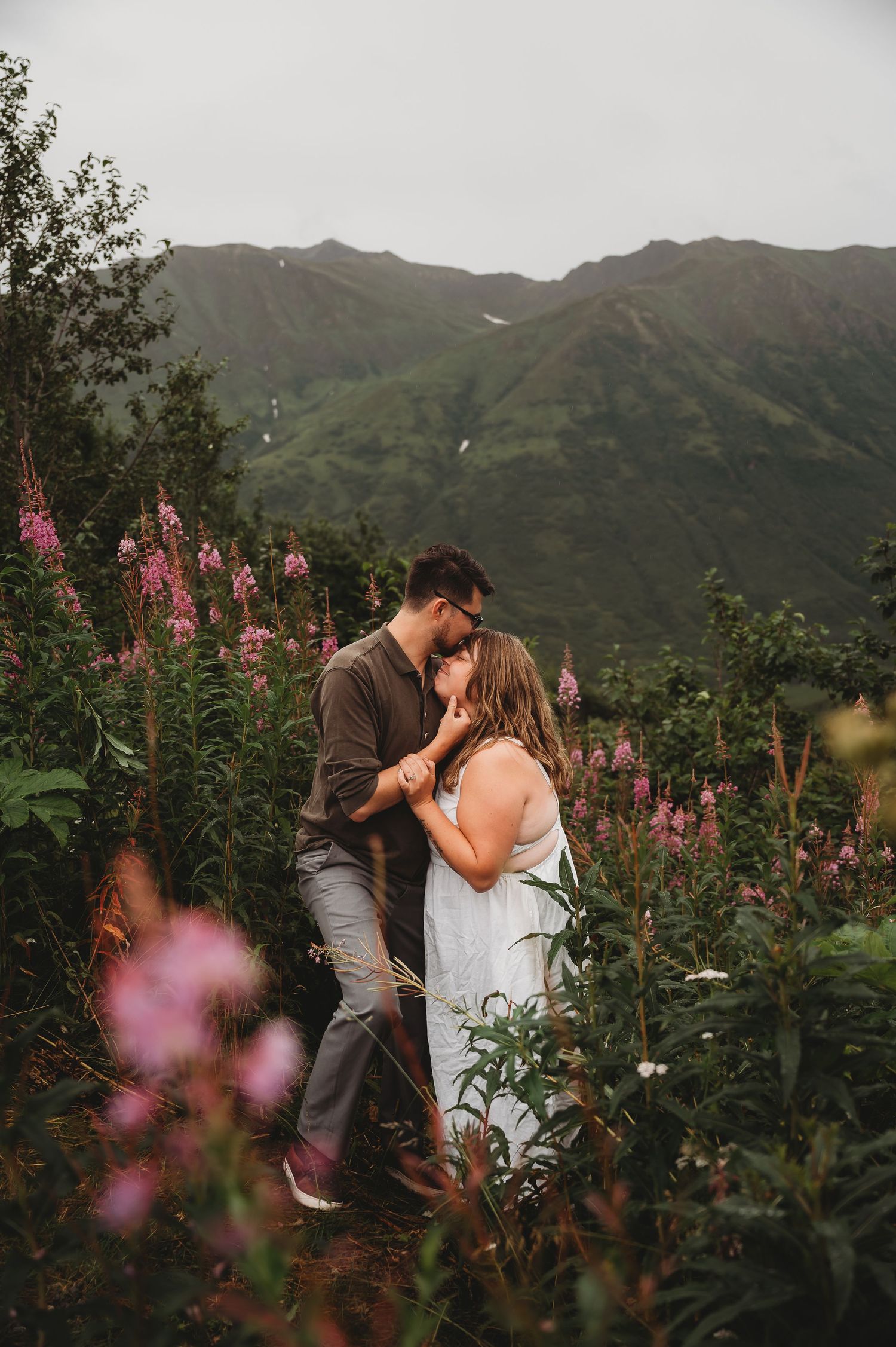 A couple shares an intimate moment among pink wildflowers with mountains in the background at sunset.
