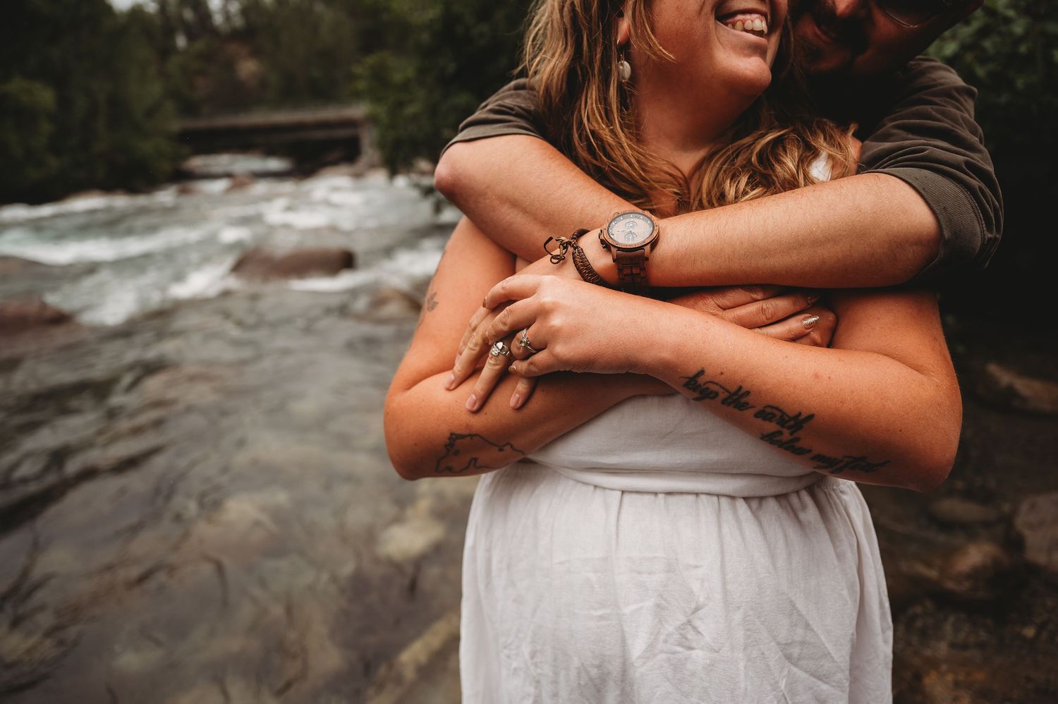 Couple embraces in loving hug by rushing river, wearing white dress and dark sweater in romantic outdoor setting.