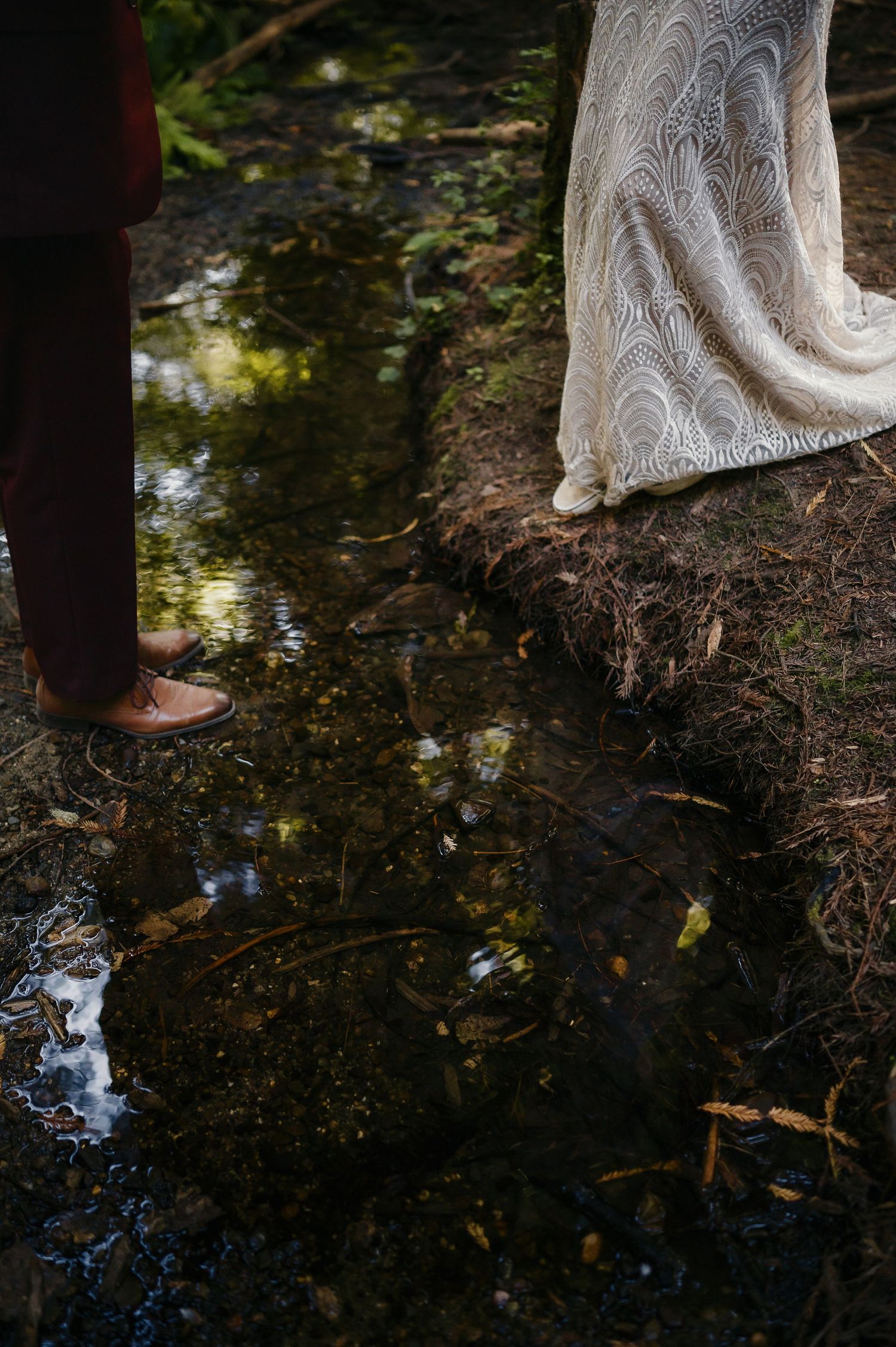 Close-up reflections in a forest puddle showing white lace dress details and forest floor.