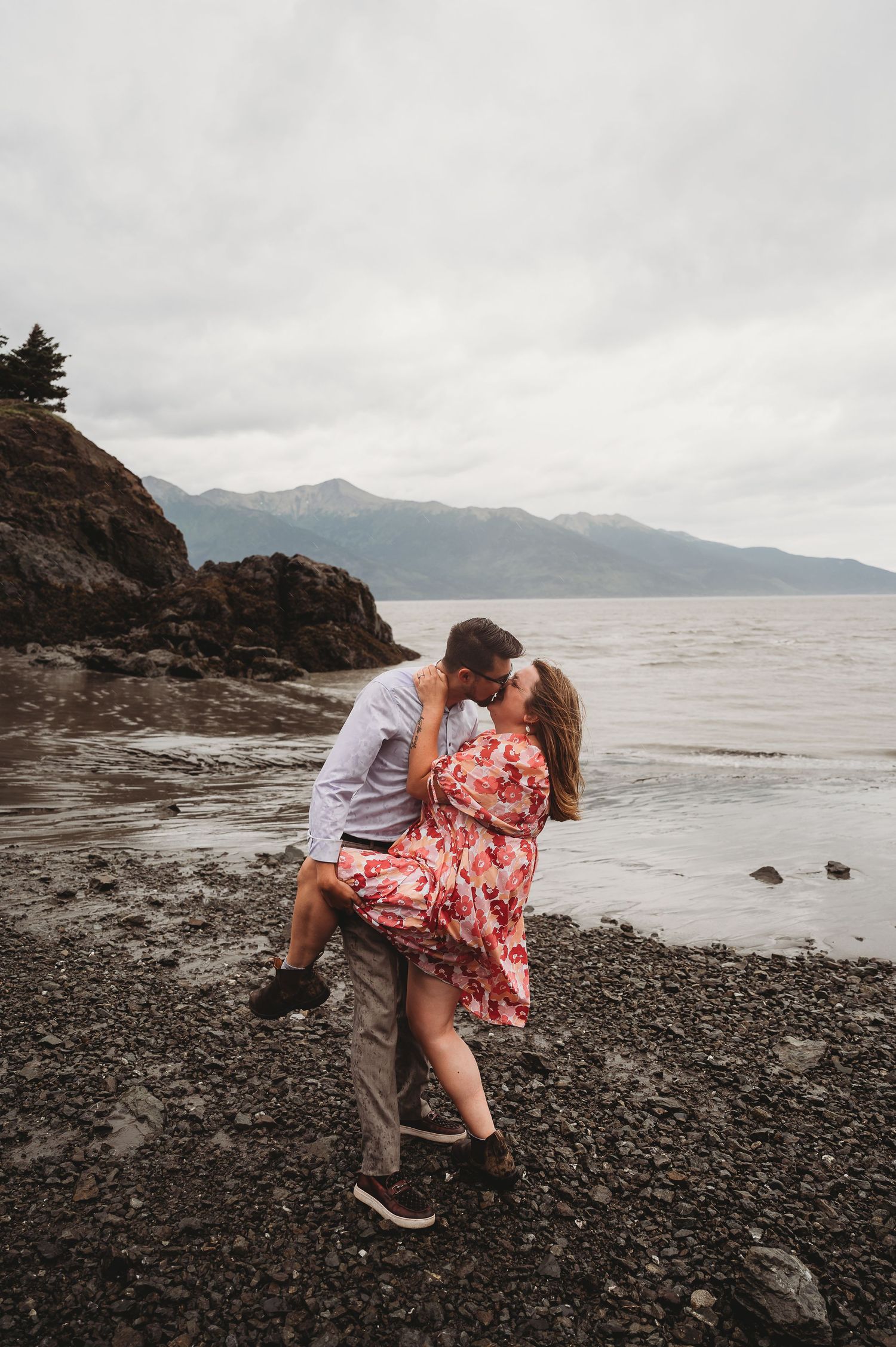 Romantic couple photography series on a rocky beach shoreline with mountains and cloudy skies in Alaska.